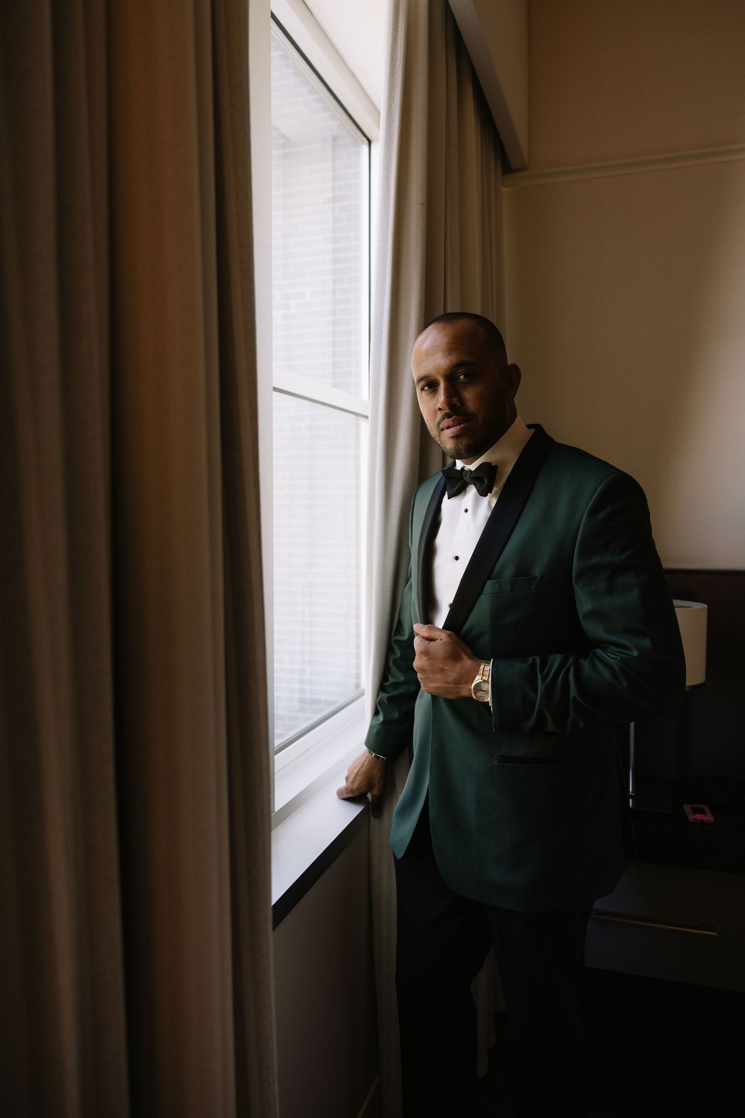 Groom in green tuxedo jacket and bow tie poses by window in dimly lit room.