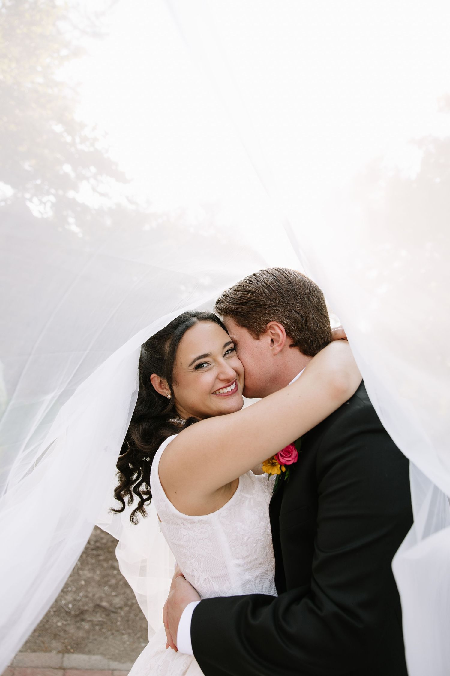 Bride and groom share a romantic moment under flowing white veil, bride smiling joyfully in white dress.