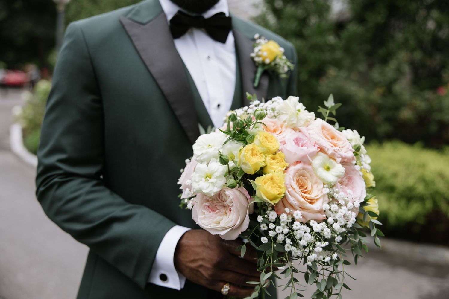 Groom in green tuxedo holding bridal bouquet of pink, yellow, and white flowers with baby's breath.