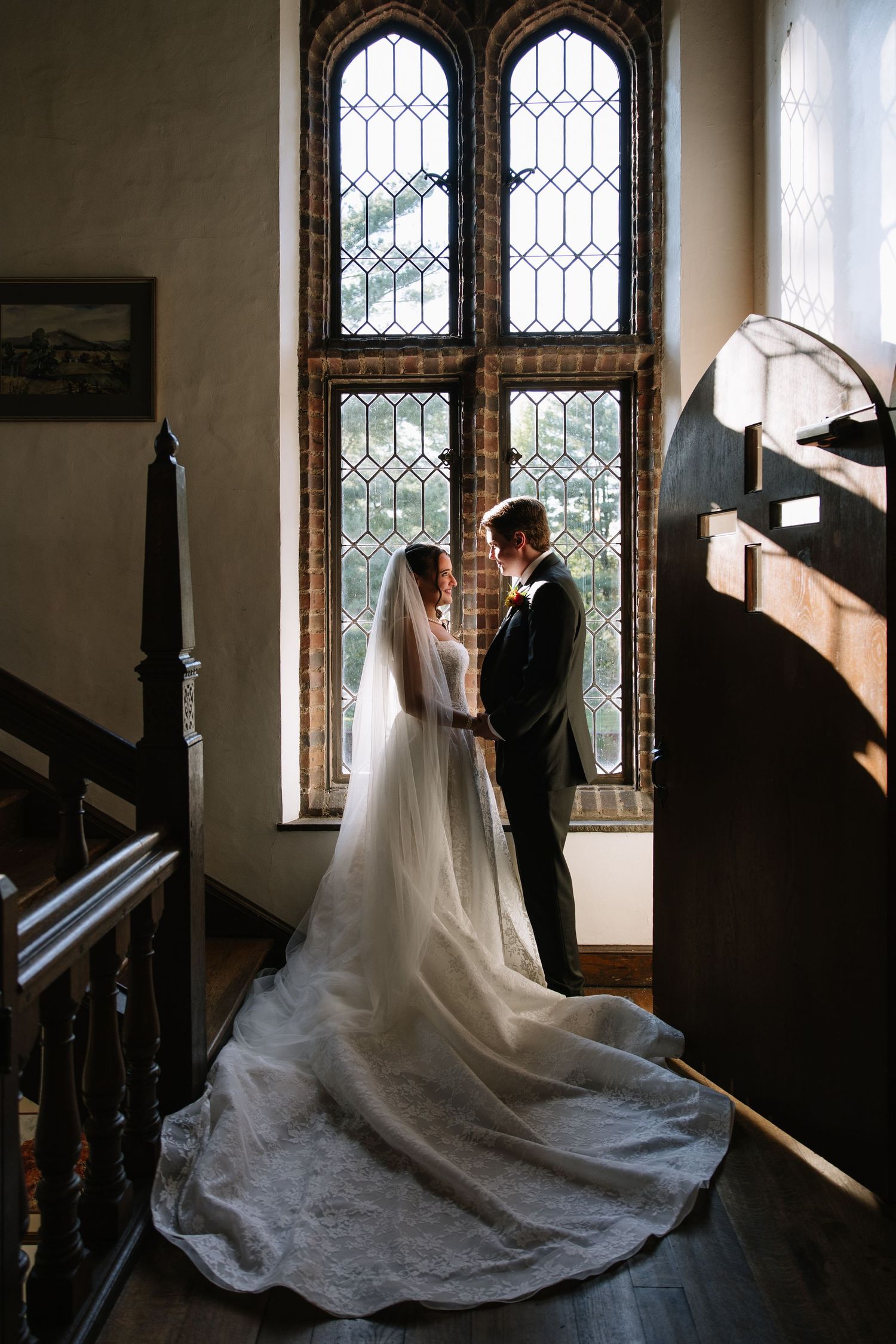 Bride and groom holding hands before gothic leaded glass windows in elegant venue staircase, wedding veil trailing on hardwood floor.