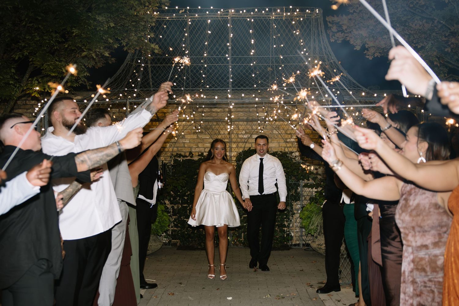 Bride and groom exit wedding reception through sparkler tunnel with guests celebrating at night.