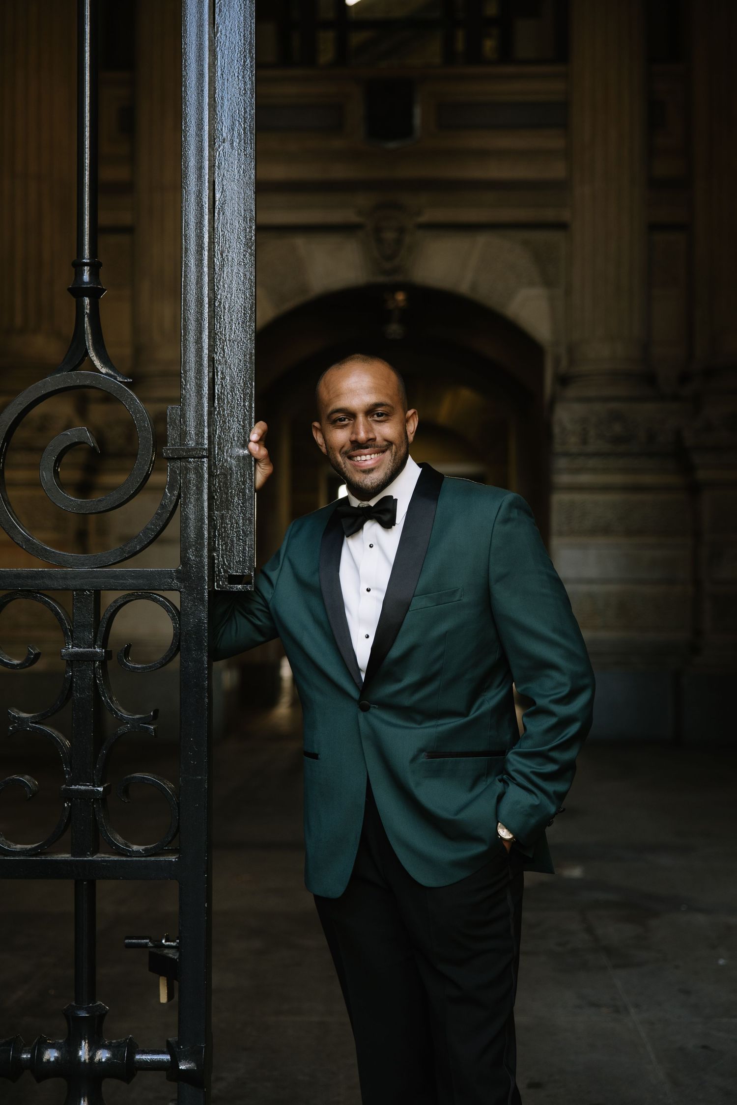 Smiling man in green tuxedo jacket and bow tie leaning against ornate iron gate in elegant stone archway.