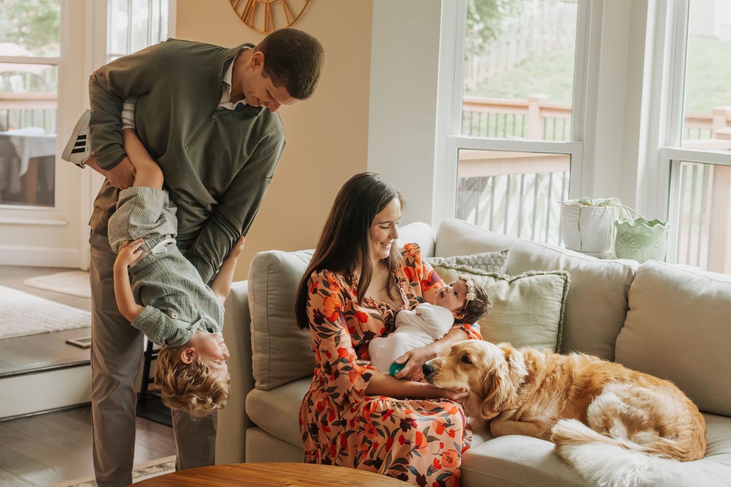 Family enjoying time at home with dad playfully lifting child, mom holding newborn on couch, and golden retriever resting nearby. St. Louis