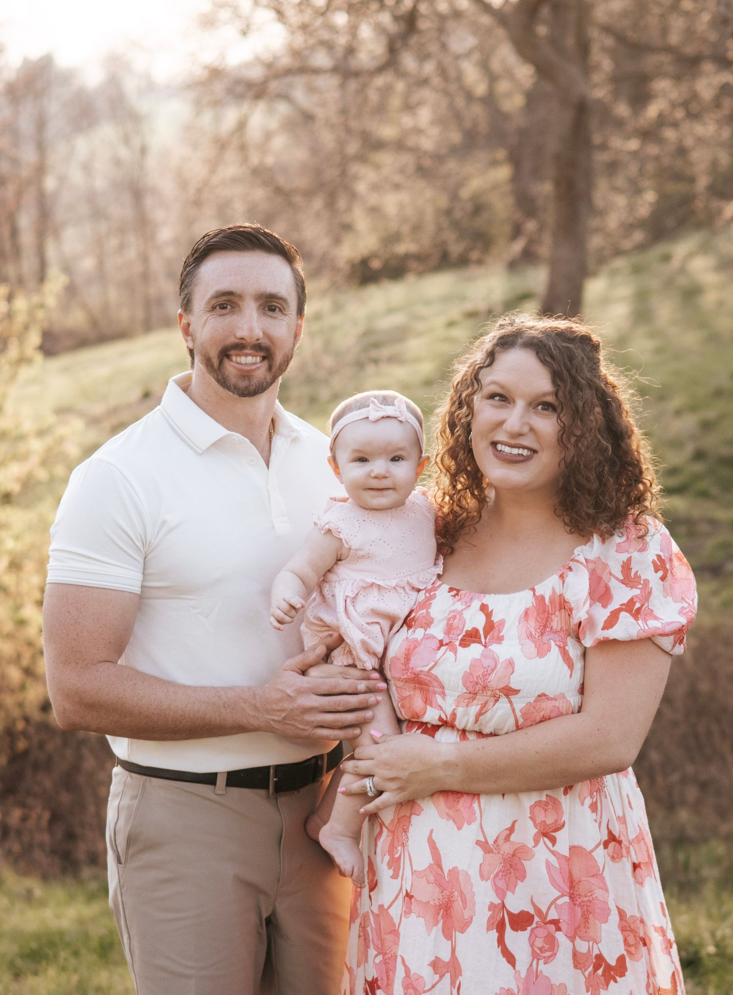 A smiling couple in a sunny outdoor setting holds their baby girl dressed in pink, surrounded by trees. Family photography, family session