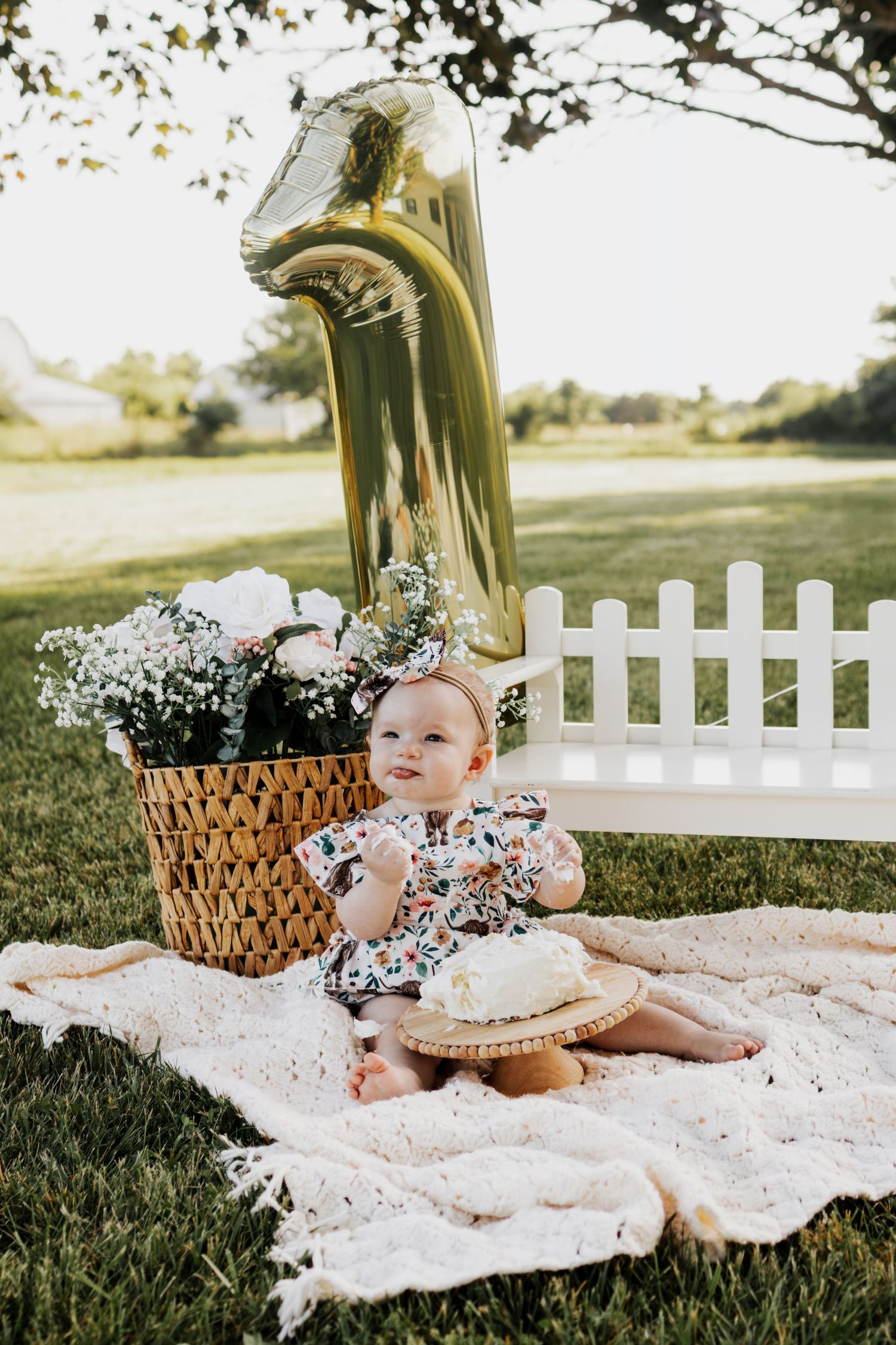 Baby girl celebrates first birthday outdoors with gold number one balloon, floral basket, and smash cake on a blanket. Troy, MO
