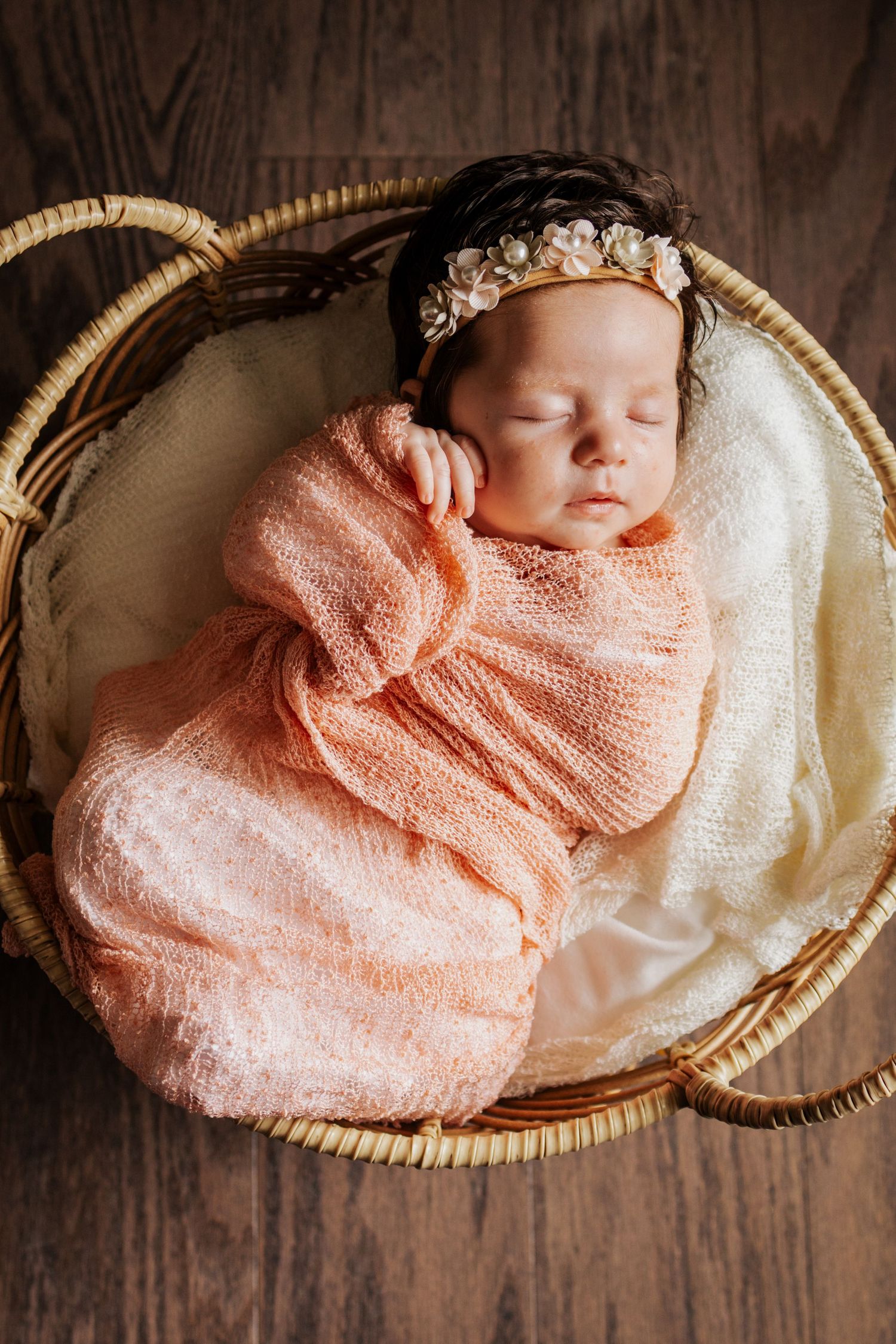 Sleeping newborn baby girl swaddled in peach wrap wearing floral headband resting in basket for her lifestyle newborn session in St. Louis,