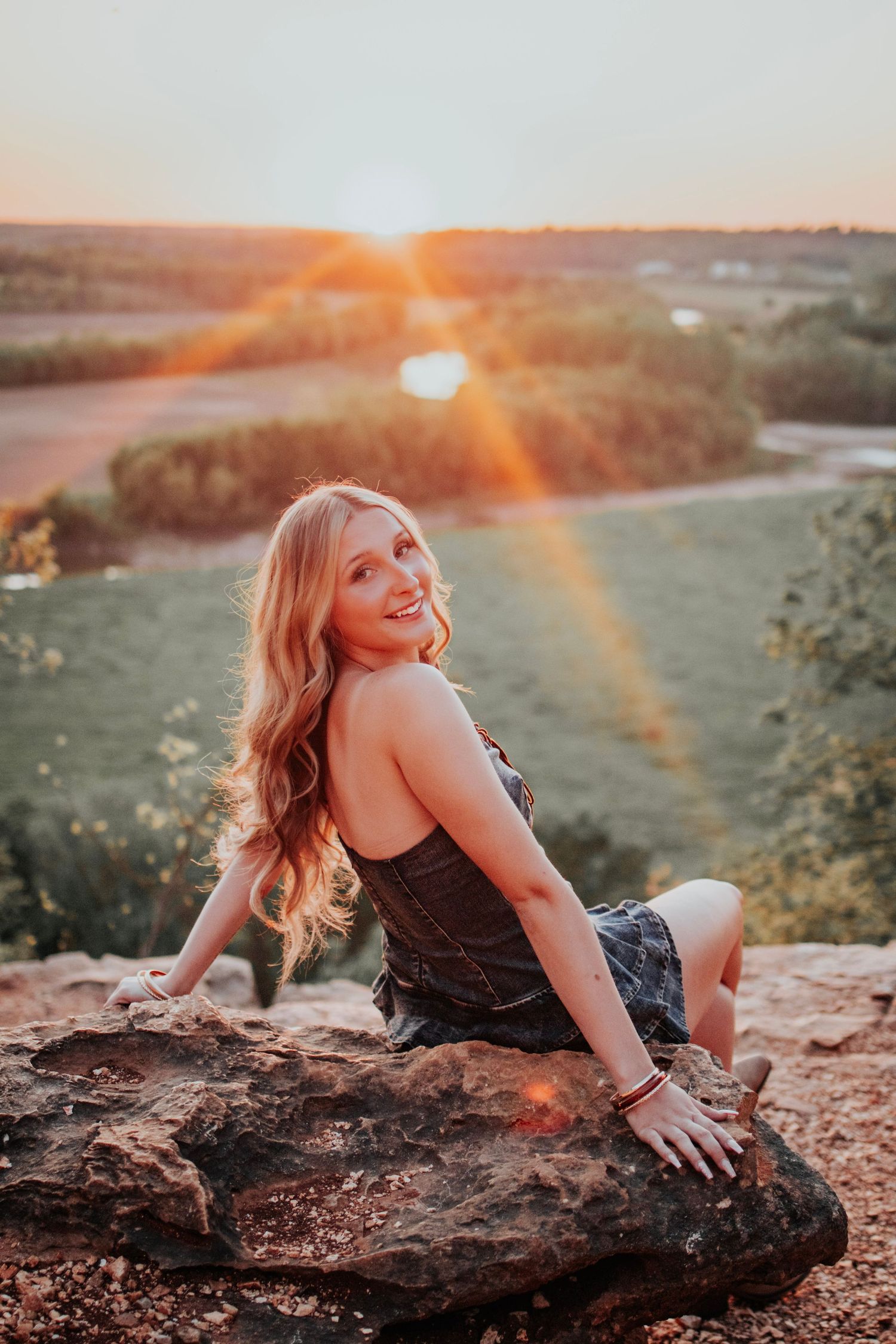 Woman sitting on rocks at sunset with golden sunlight and green landscape in background during her senior photo session in Troy, MO