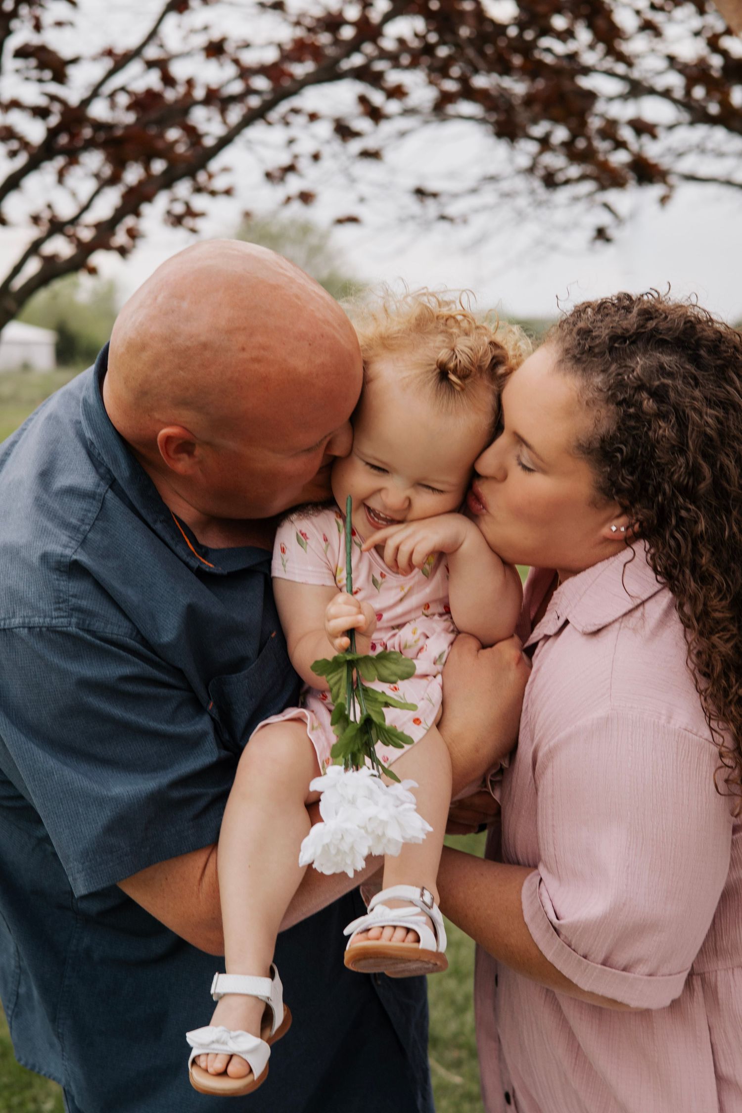 Parents kissing their laughing toddler girl on cheeks outdoors, child holding white flowers, family portrait in Troy, MO.