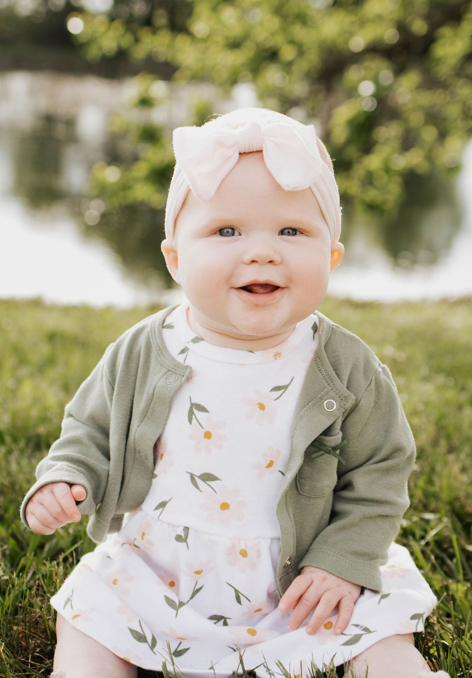 Infant girl in a olive green cardigan and floral white dress smiling at the camera in Troy, MO during family session
