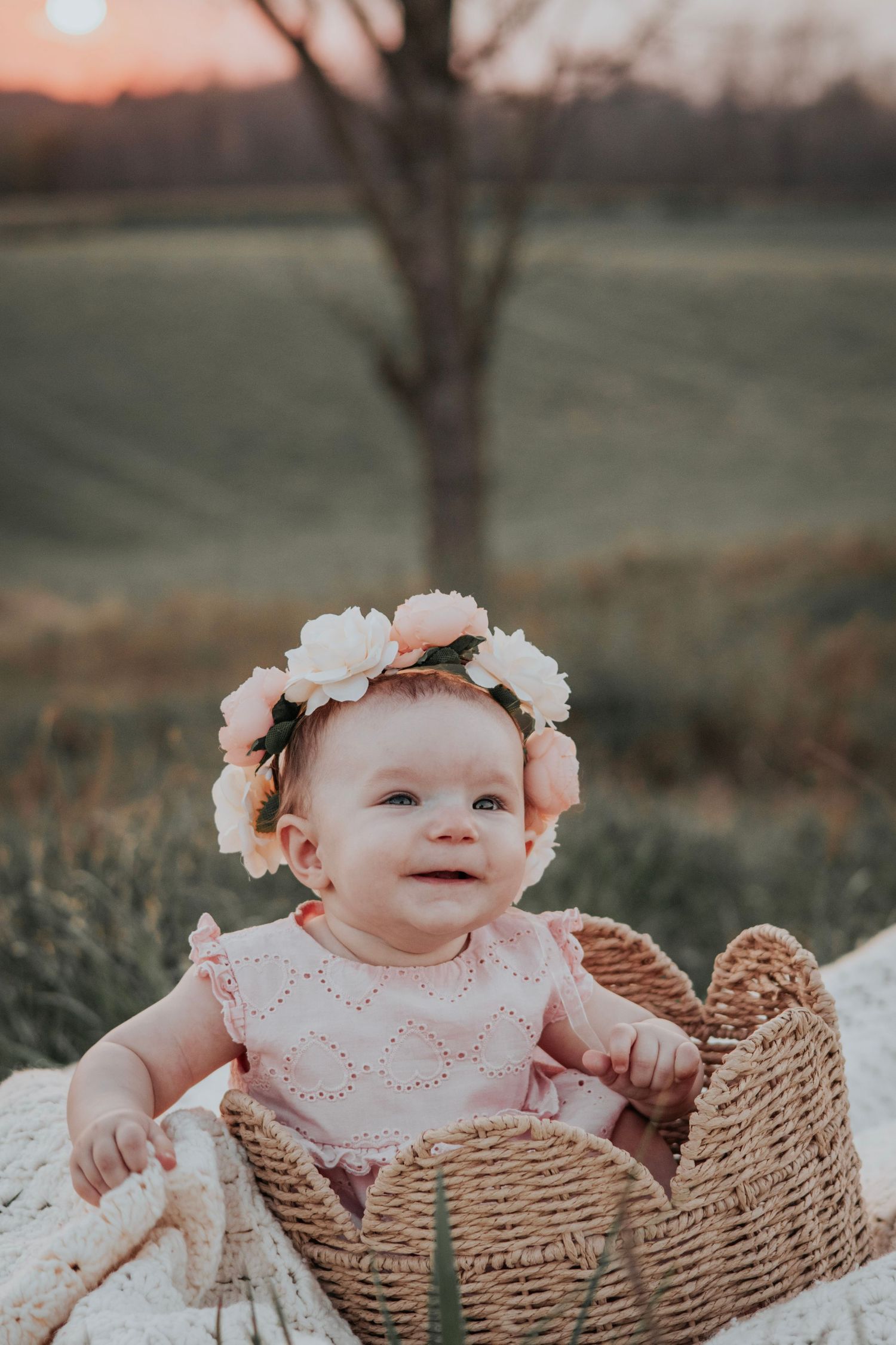 Infant girl sitting in a scallop trimmed basket with a orange/pink sunset in the background, baby wearing flower crown. Family session