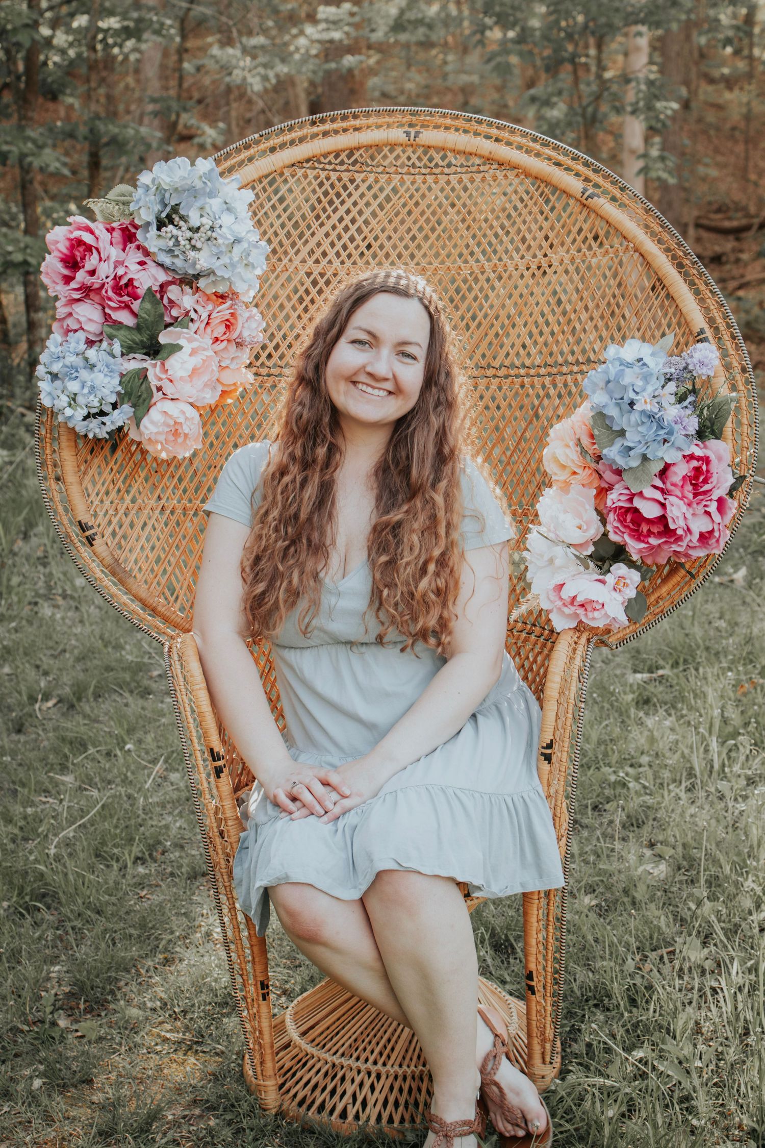 Young woman sits in a peacock chair decorated with spring florals in a wooded area
