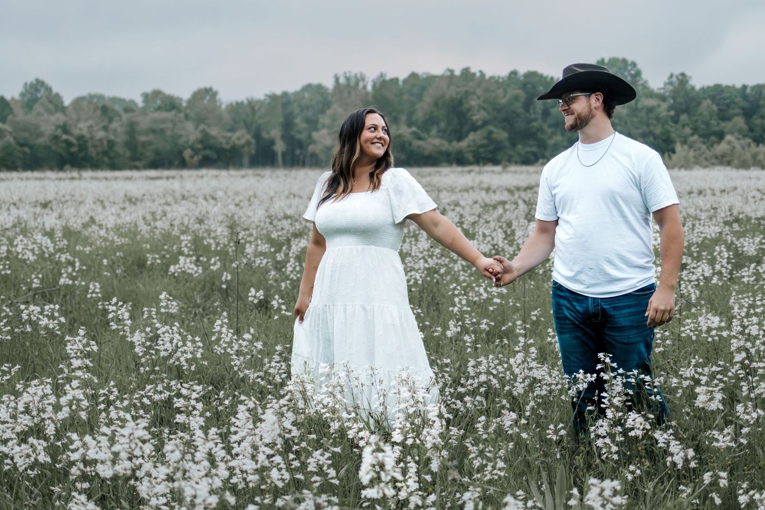Couple holding hands in a white wildflower field, woman in white dress, man in cowboy hat and jeans. Engagement session, Foristell, MO