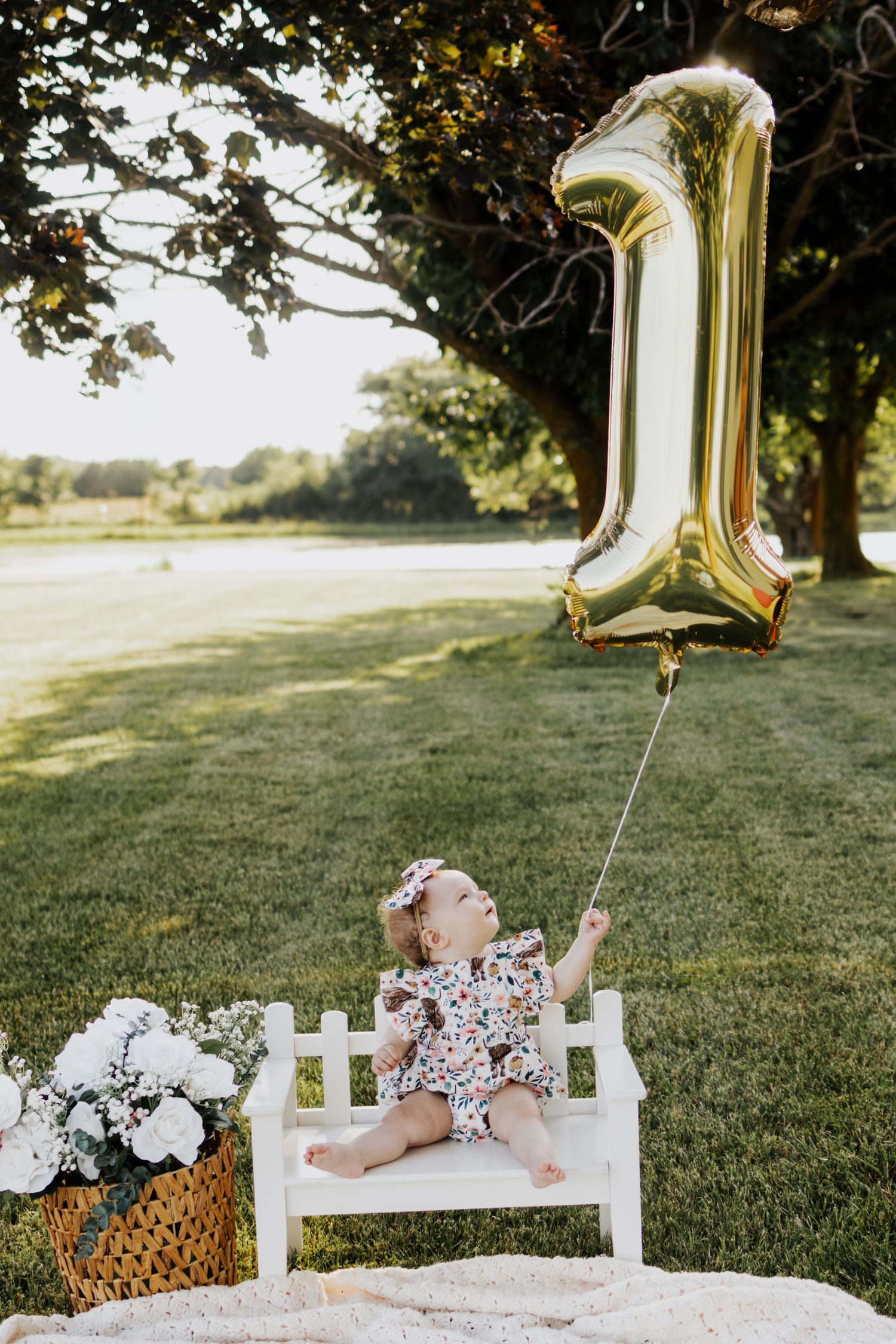 Baby girl in floral outfit sits on white bench holding gold number 1 balloon for first birthday celebration outdoors in Troy, MO.