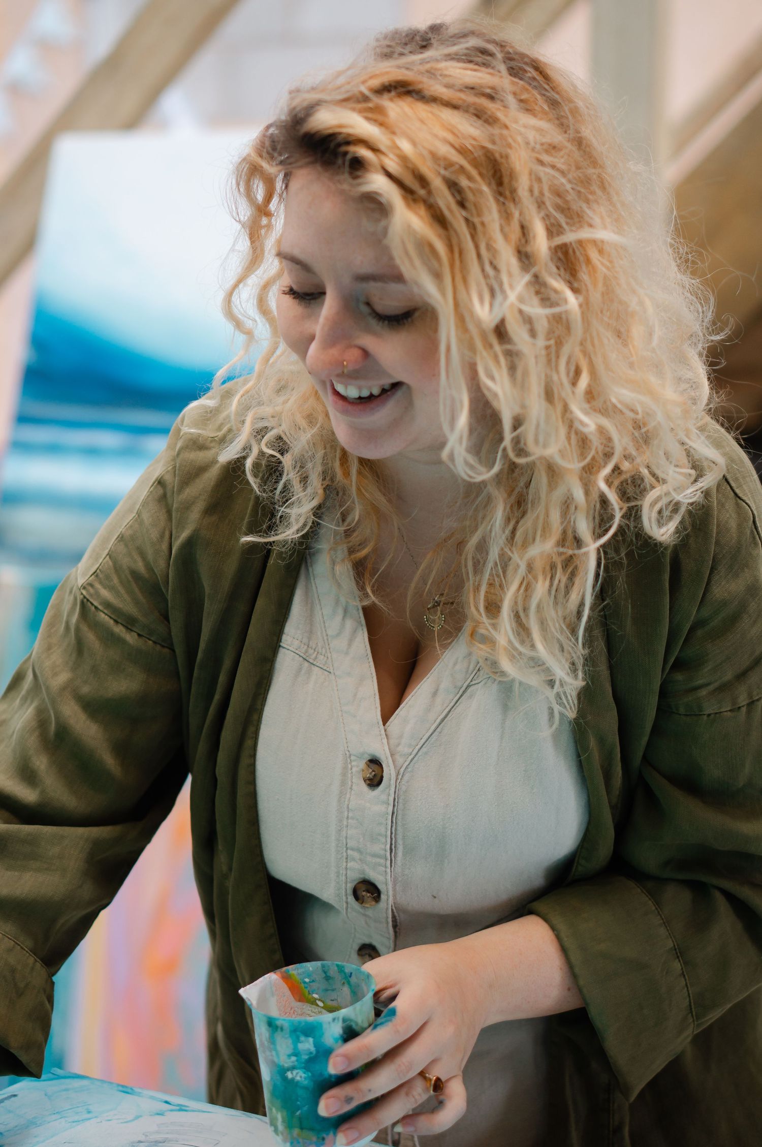 Georgia Morgan, Braunton based artist, with curly blonde hair wearing a green jacket holds a colorful cup painting in her studio.