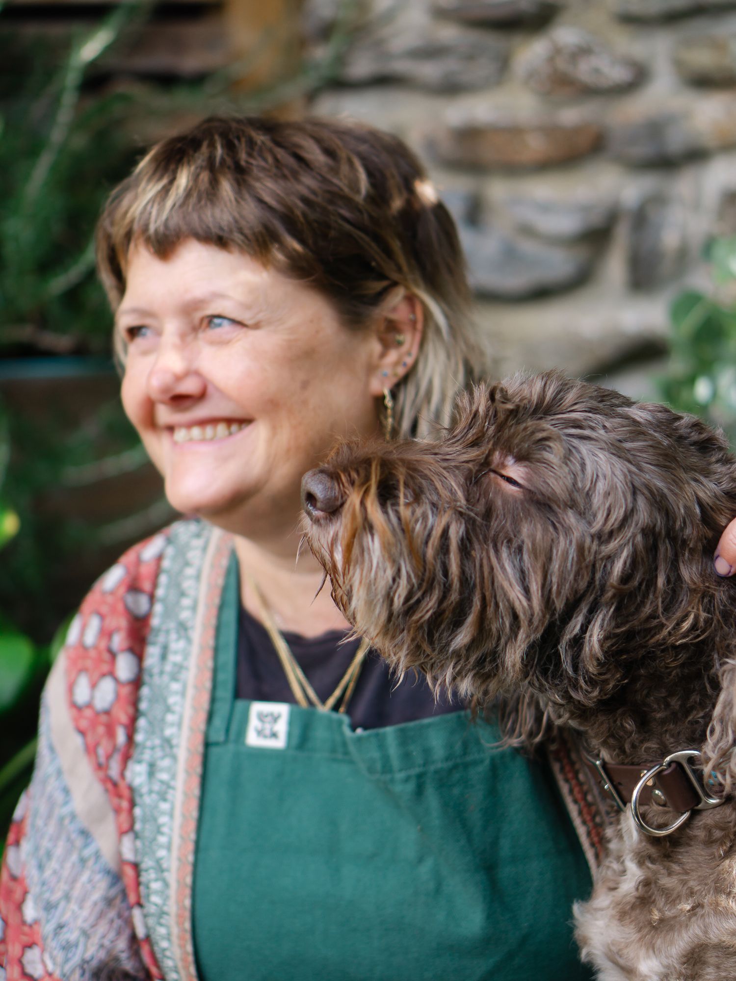 Newton Abbot based tattoo artist Linda Dinata in green apron with a curly brown dog resting on her shoulder outdoors near a stone wall.