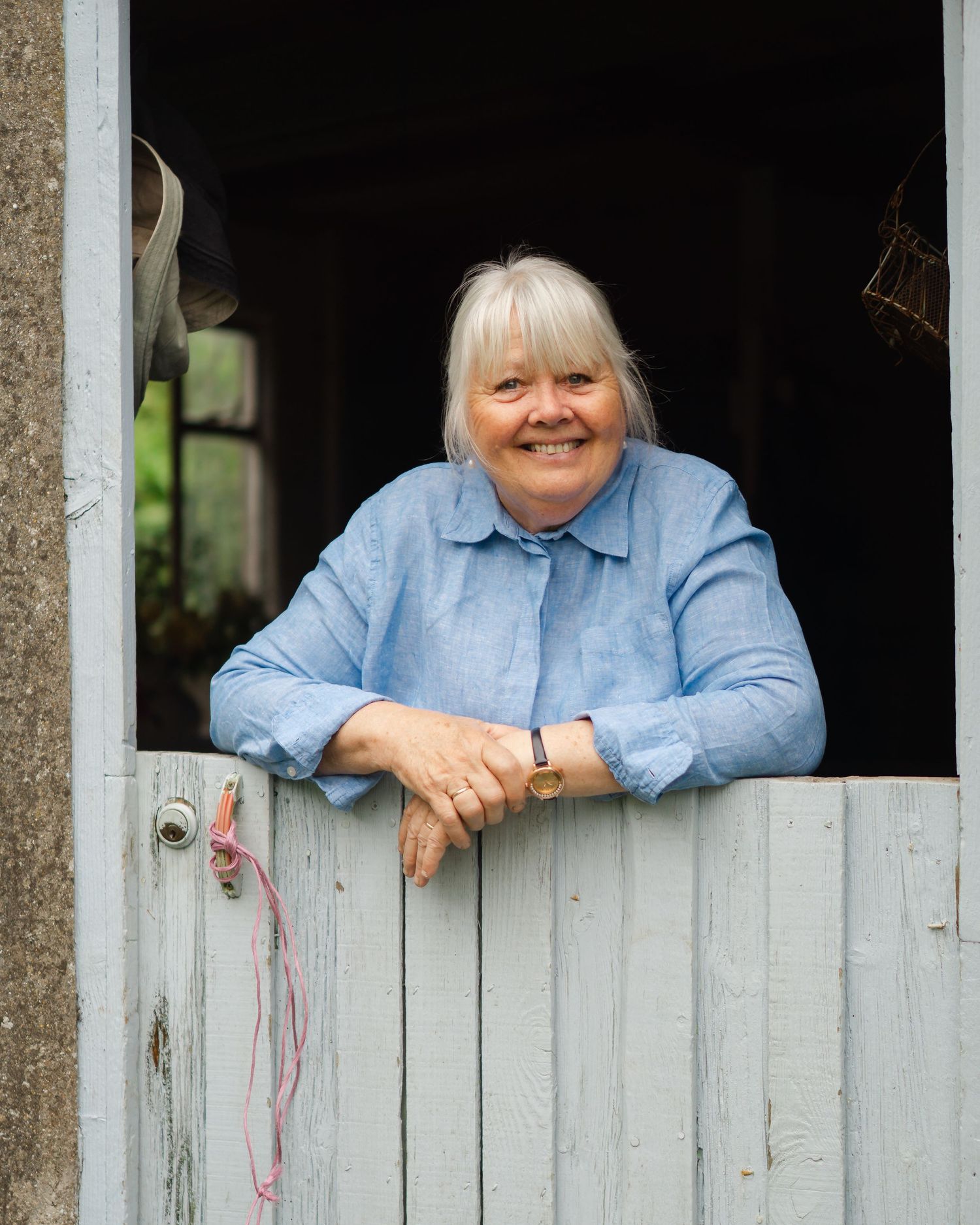 Smiling older woman with gray hair leaning on a rustic wooden stable door wearing a blue linen shirt.