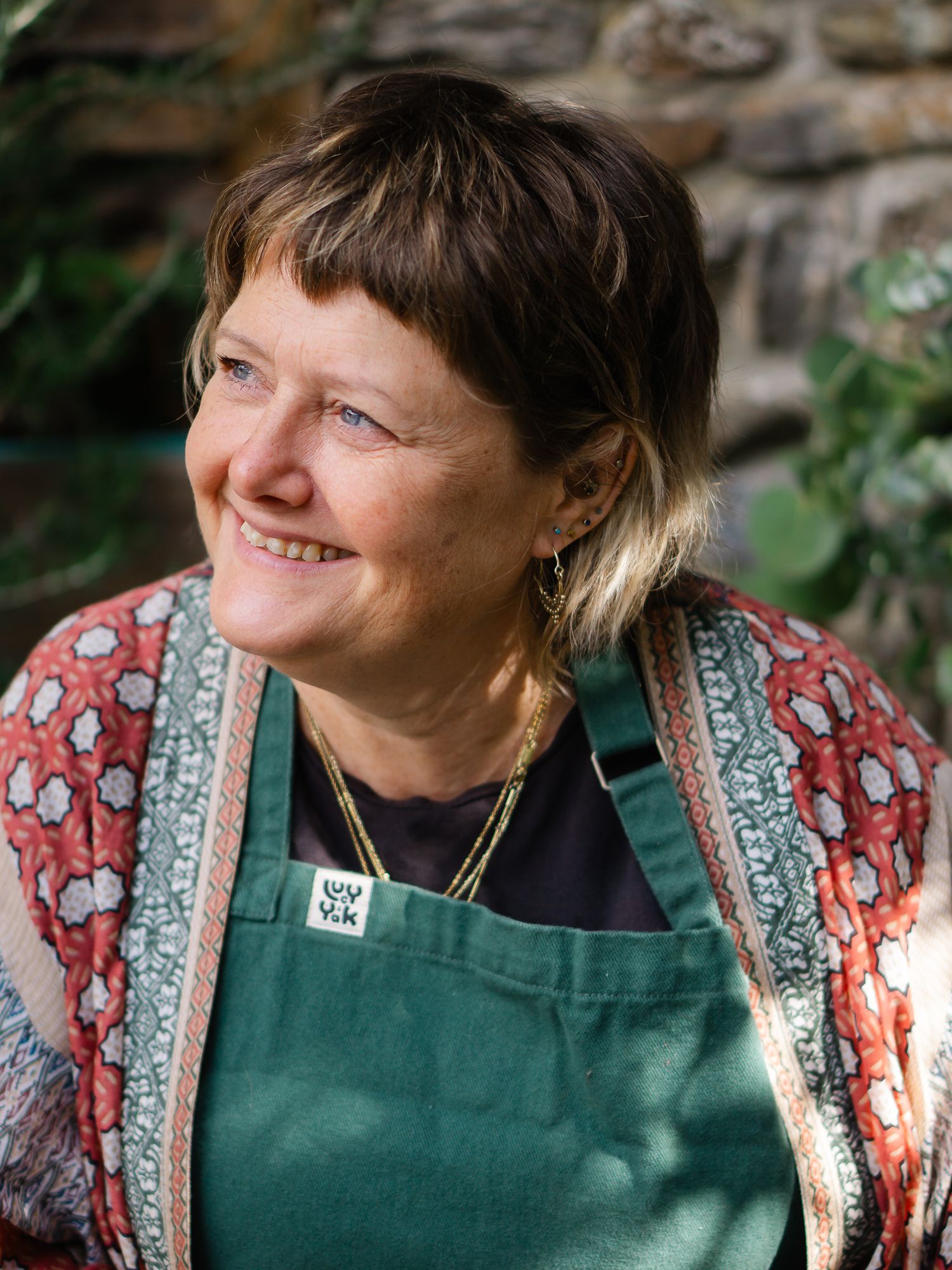 Smiling middle-aged woman with short brown hair wearing a green apron and floral jacket outdoors.