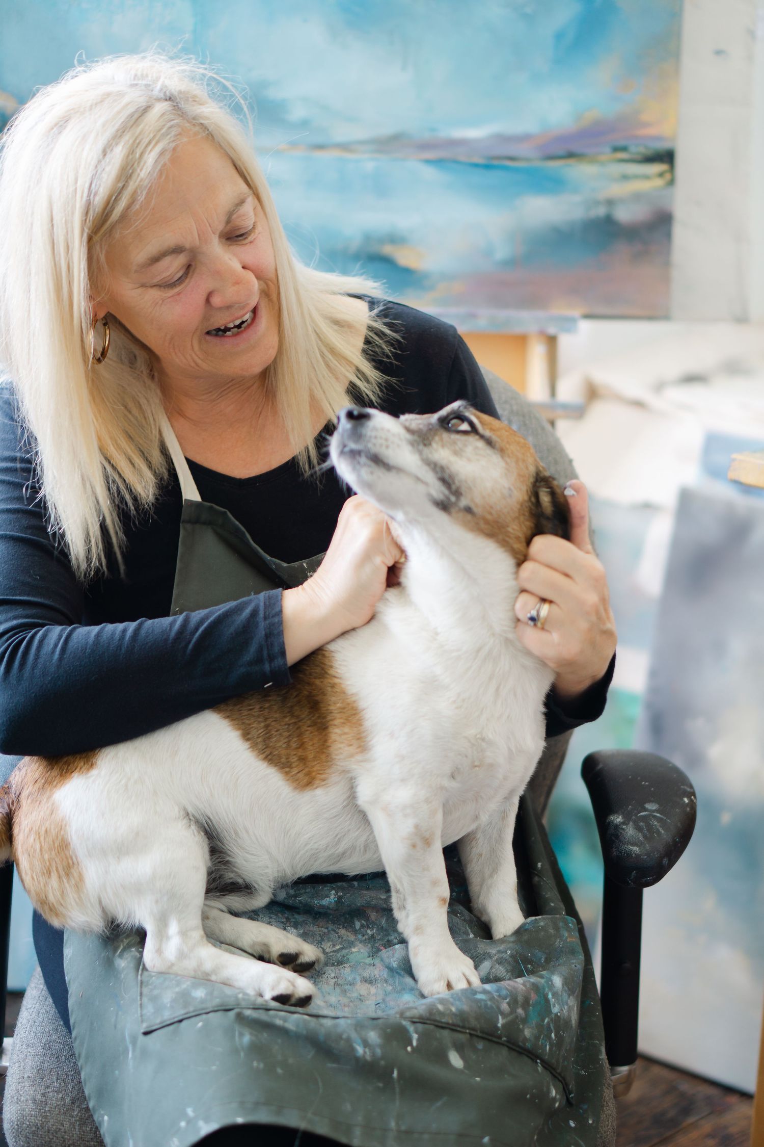 Smiling Jo Payne, in her art studio petting a small Jack Russell terrier dog sitting on a paint-splattered stool.