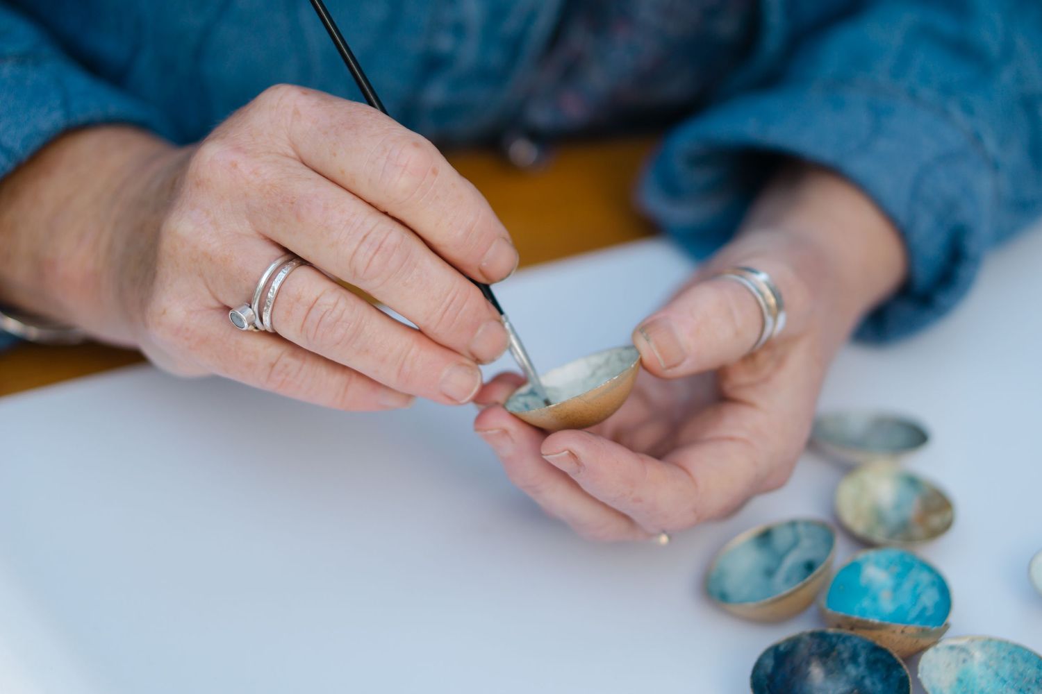 Close-up of hands painting a small stone with a fine brush, with colorful painted eggshells displayed on a white surface.