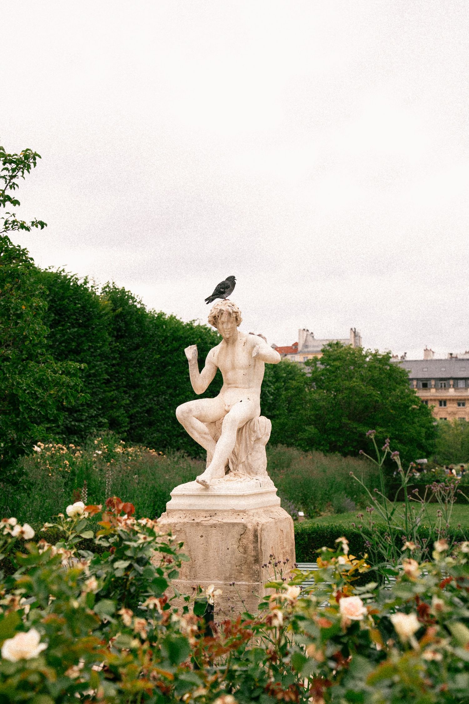 A white classical marble statue with a crow perched on top, surrounded by blooming flowers and lush green trees in a garden.