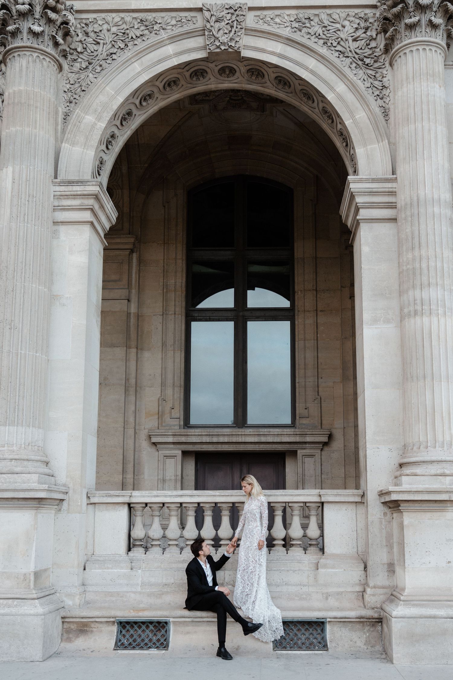Bride and groom pose on ornate stone steps beneath a grand arch at the Louvre in Paris, France.