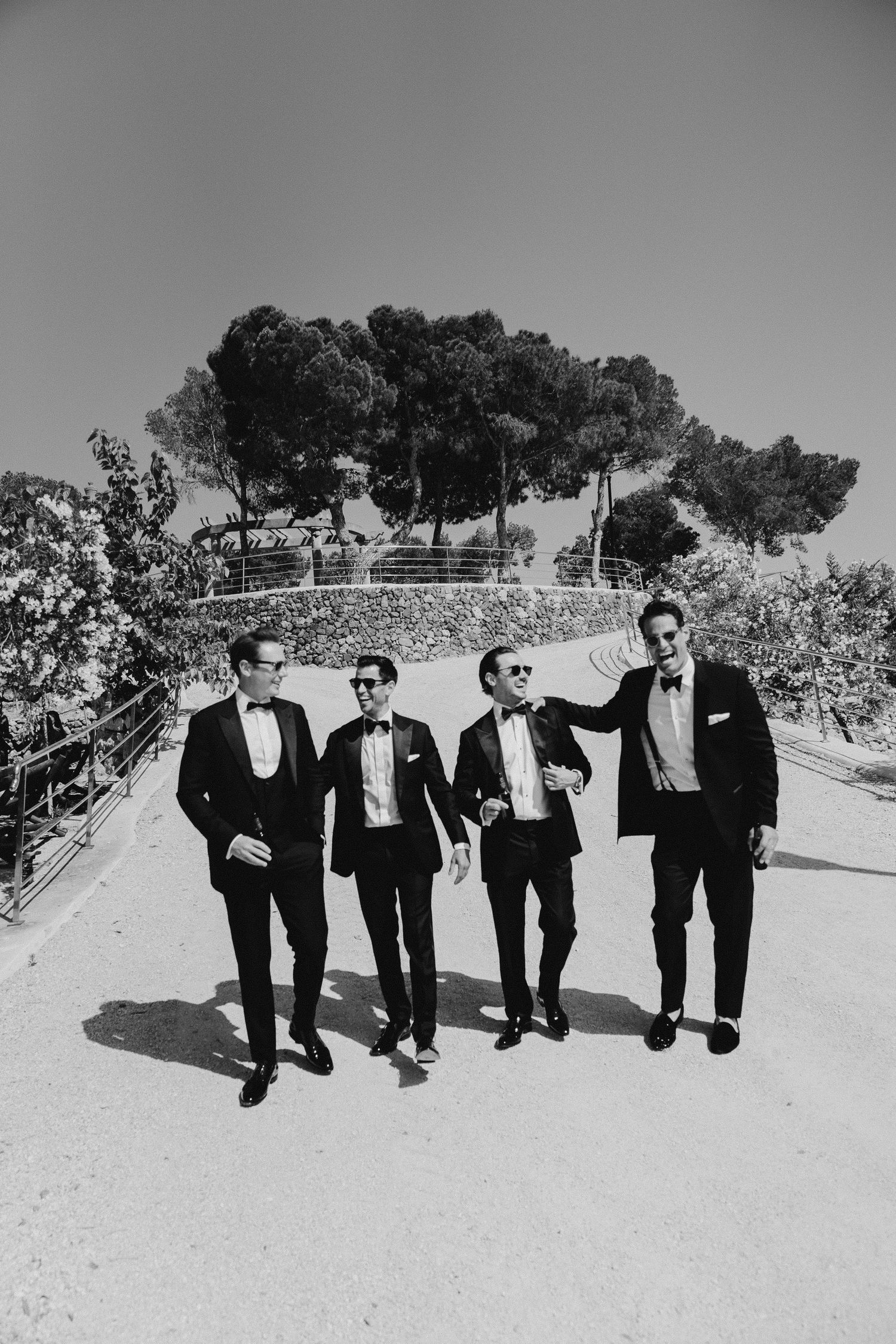 Four groomsmen in black tuxedos walking confidently on a sunny path in black and white photo.