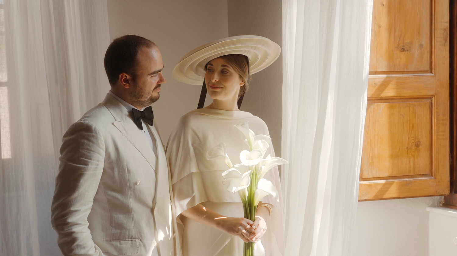 Groom in white suit stands beside bride wearing a wide-brim hat holding white calla lilies near a window.