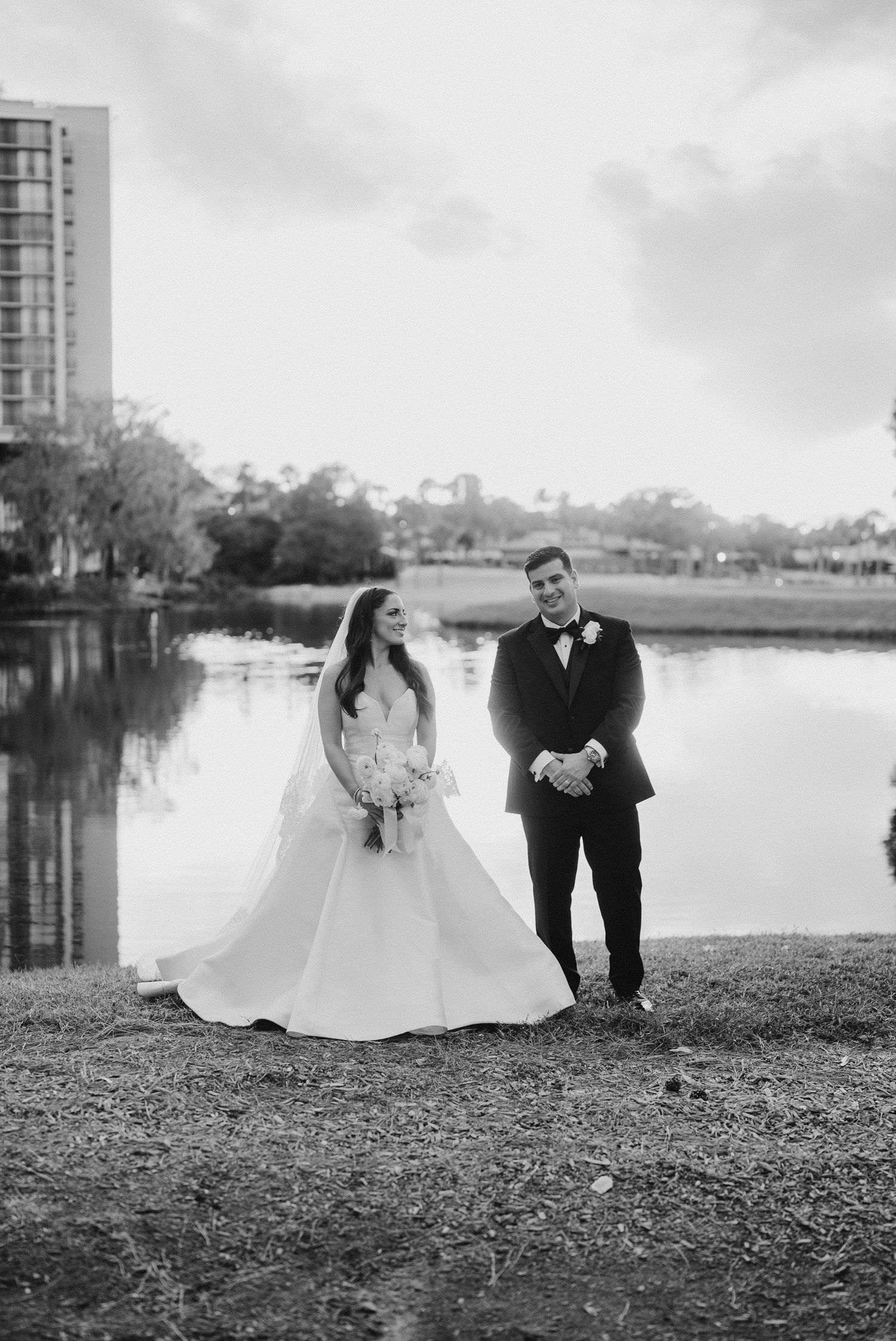 Black and white photo of bride in ball gown and groom posing by a peaceful lake.