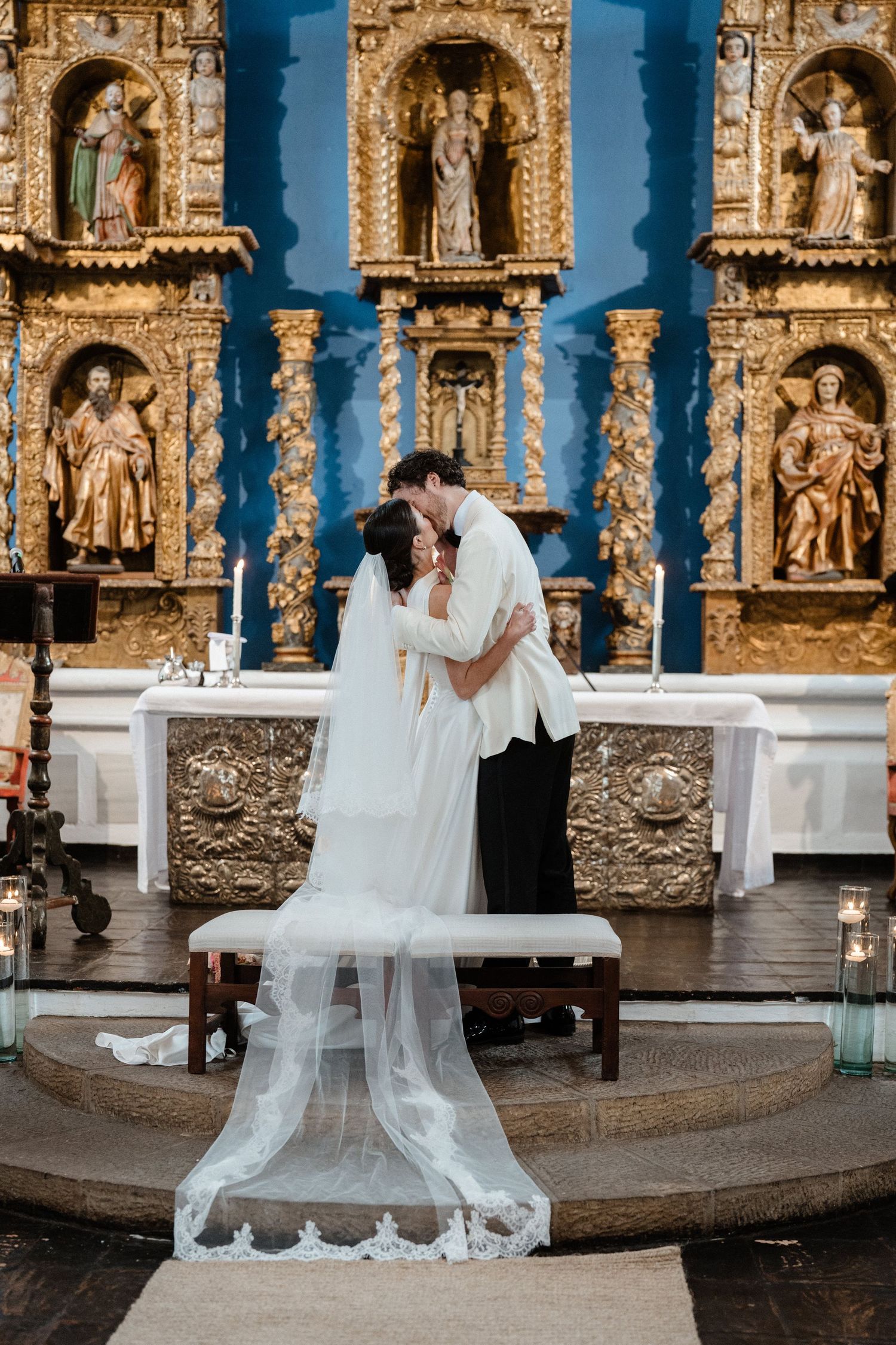 Bride and groom share first kiss at altar in front of ornate golden retablo in historic church.
