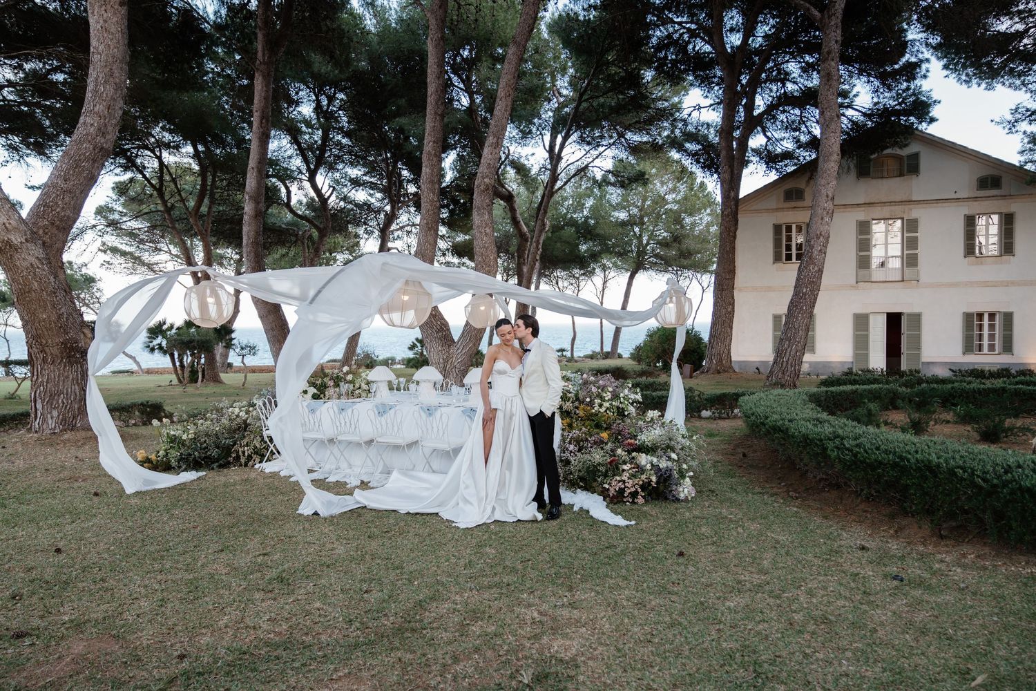 Bride and groom pose under elegant white wedding arch in lush garden with trees and villa backdrop.