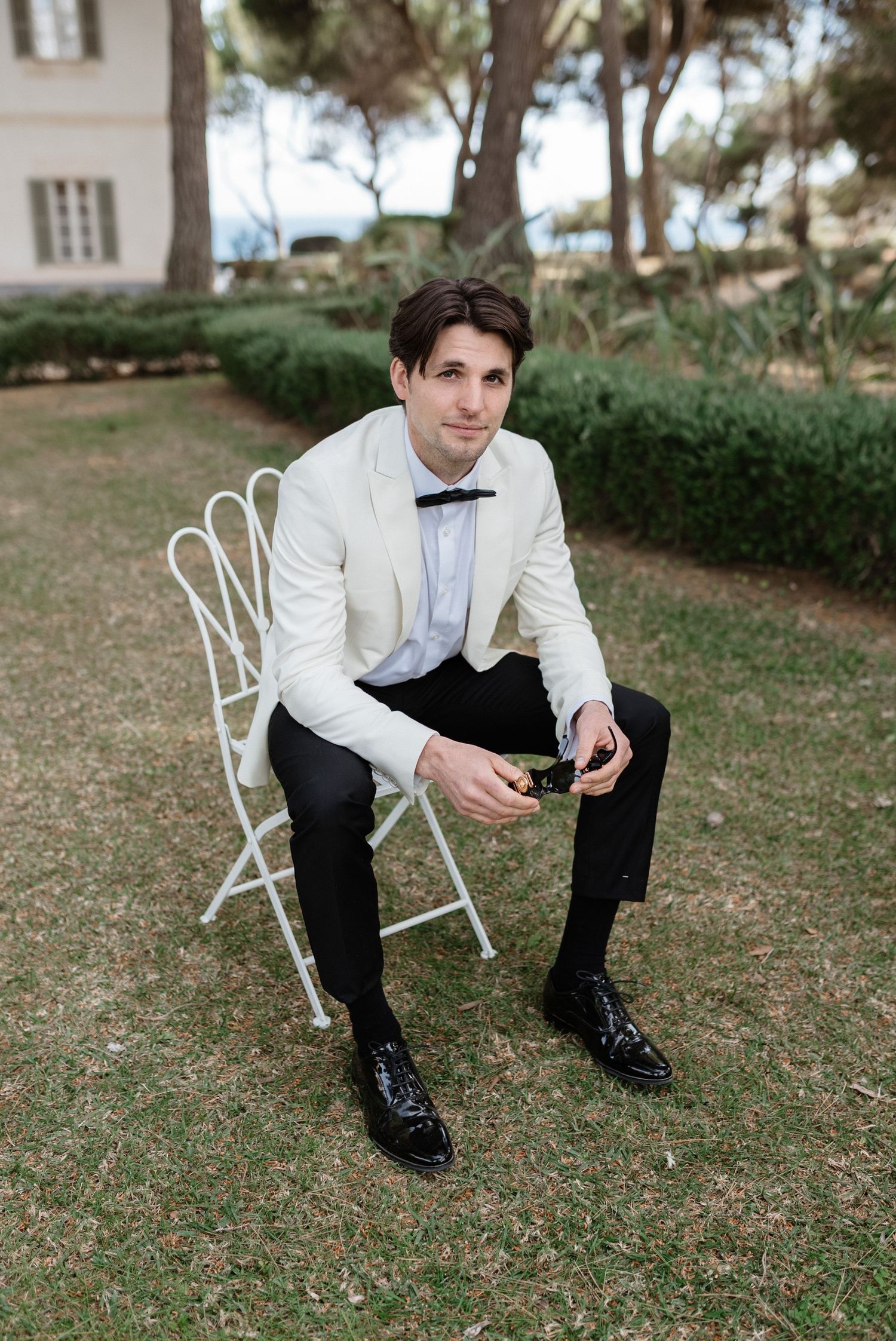 Dapper groom in cream tuxedo jacket and bow tie seated on white chair in garden setting.