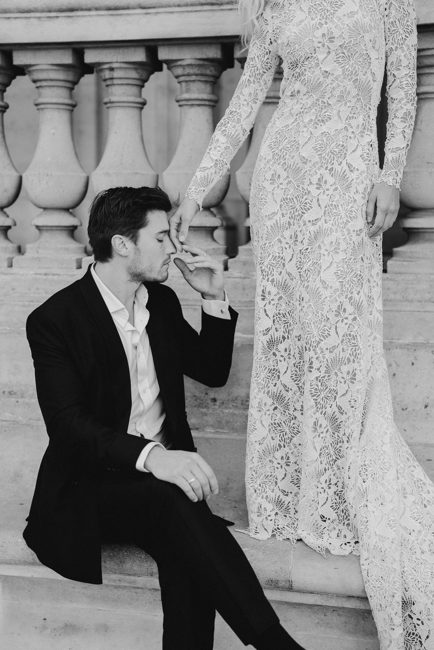 Groom in black suit kisses bride's hand while seated on stone steps, bride in lace wedding dress, black and white photo.