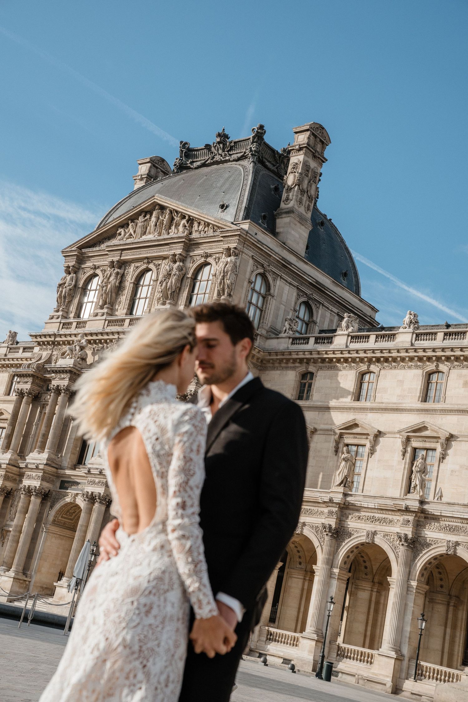Bride in backless lace gown and groom in black suit embrace before the iconic Louvre Museum in Paris.