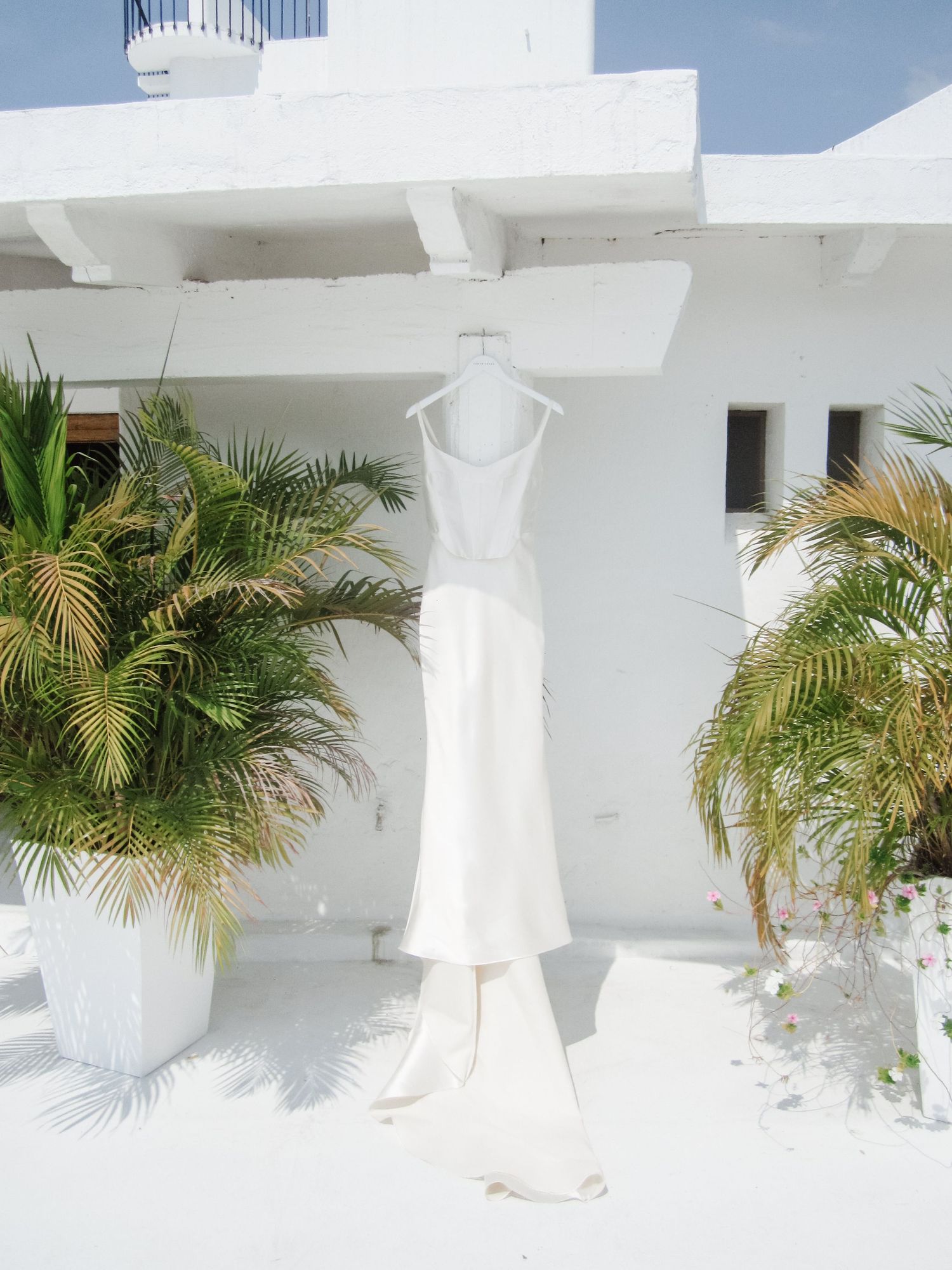 White wedding dress hanging on a hanger outside a tropical white villa with palm trees.