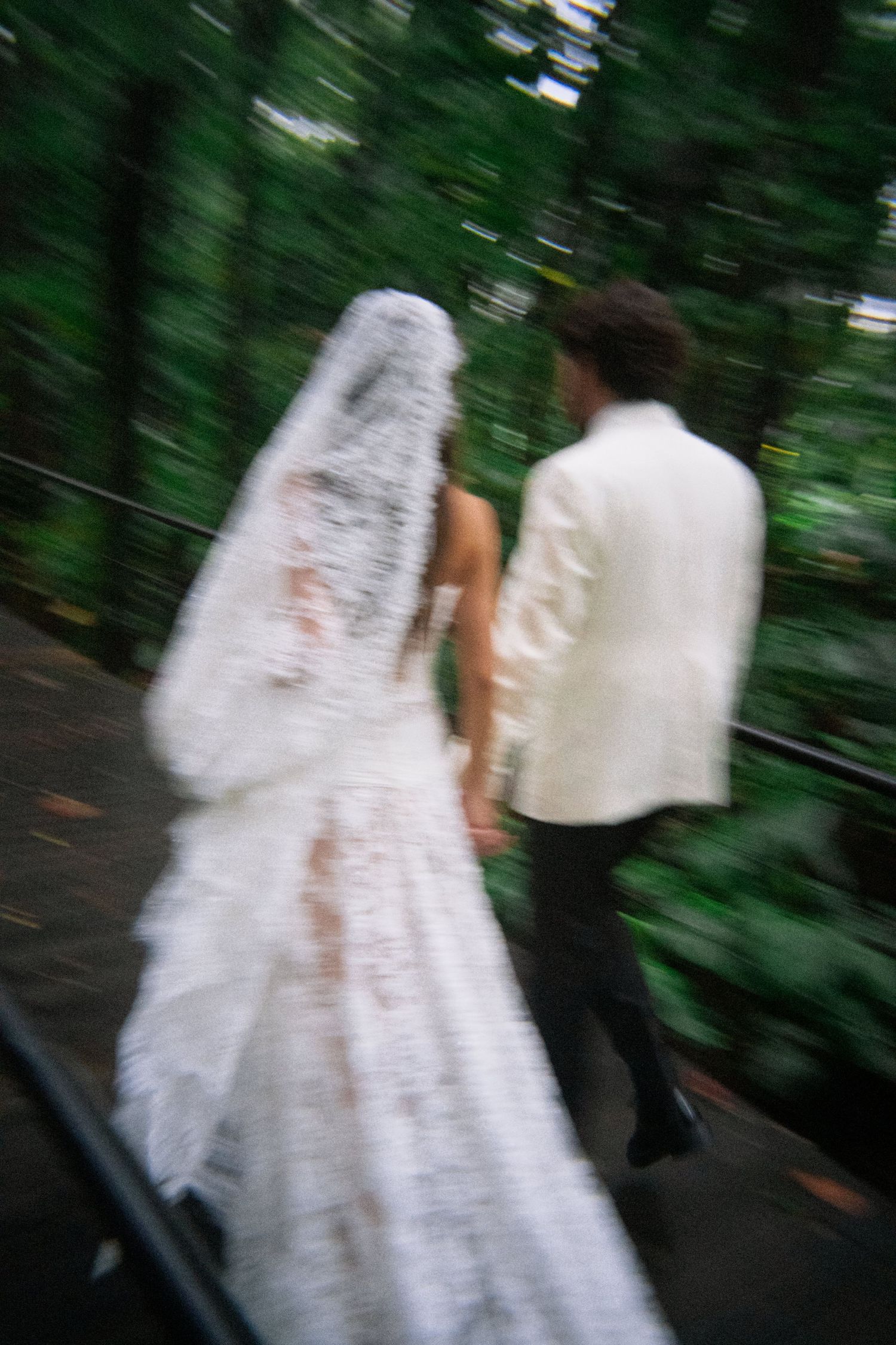 Blurred motion photo of bride and groom walking hand in hand through a lush green forest.