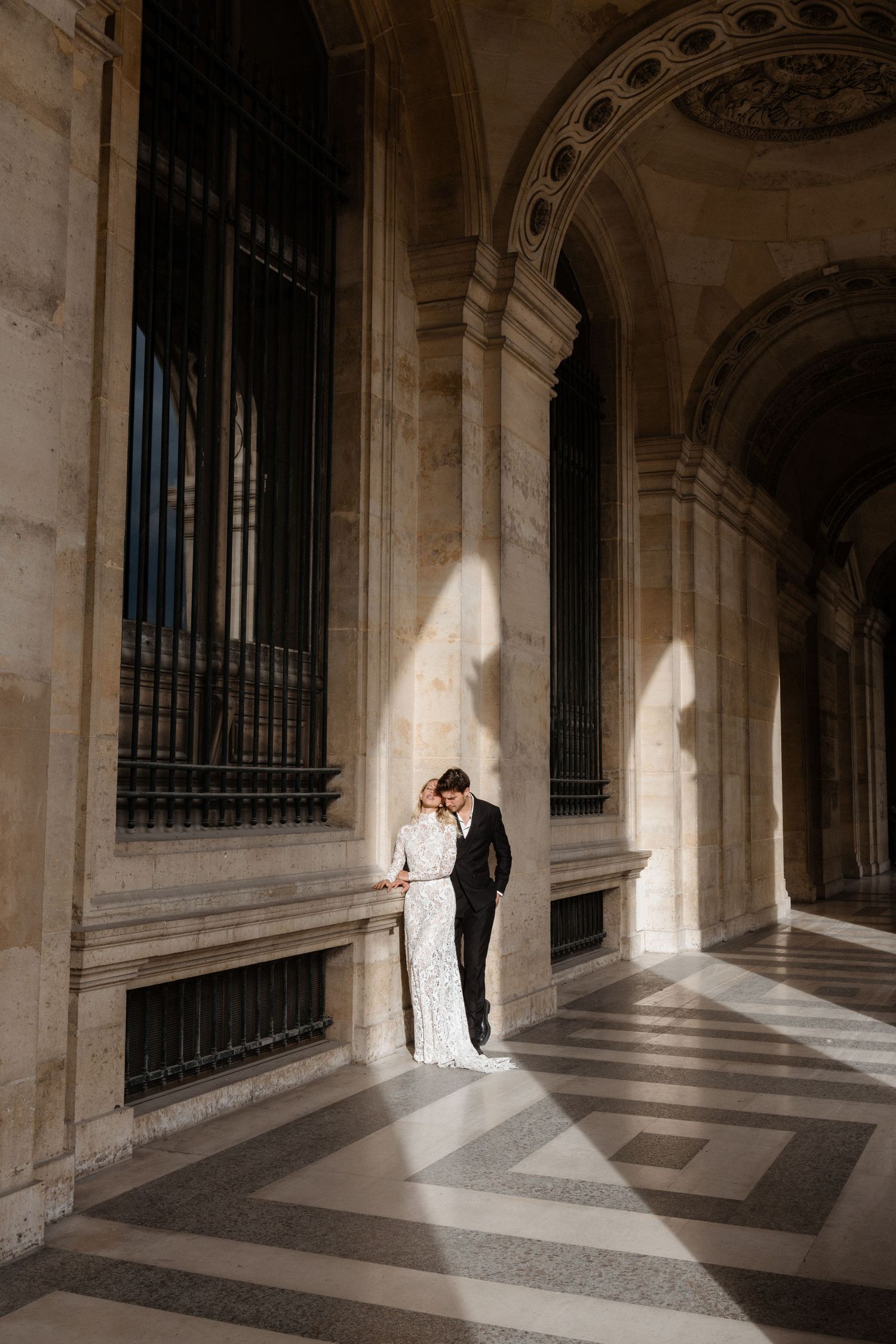 Bride and groom sharing intimate moment in sunlit ornate marble hallway of classic European building.