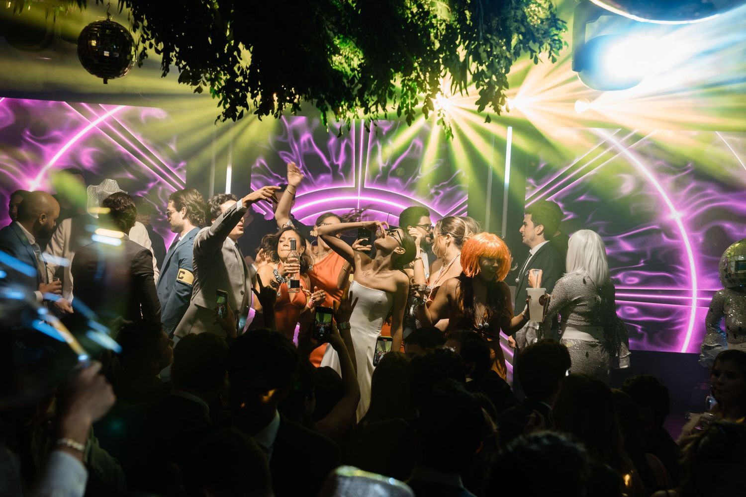 Joyful wedding reception crowd dancing under colorful purple and green disco lights outdoors at night.