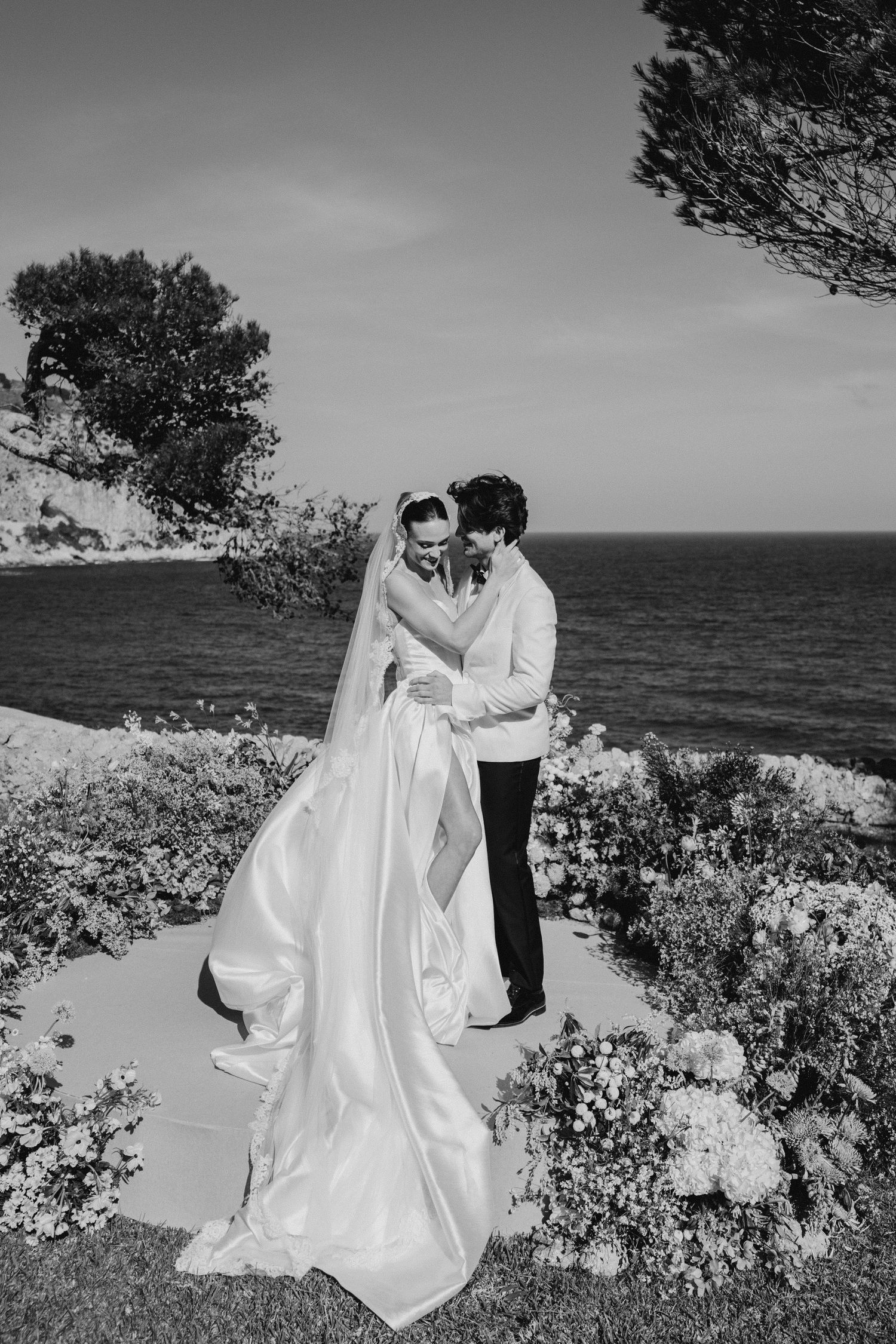 Black and white portrait of bride and groom embracing by the sea with floral arrangements nearby.