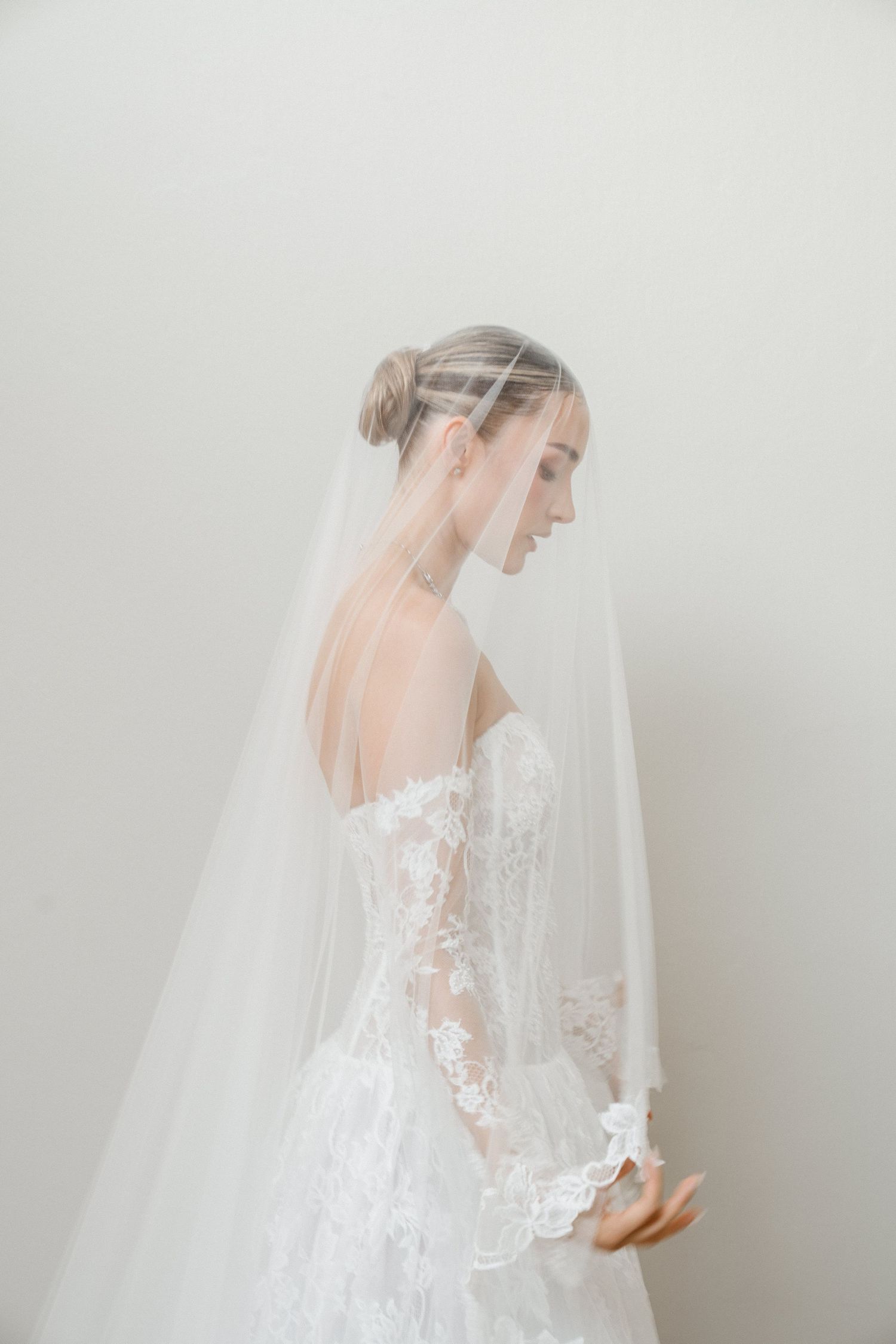 Bride in strapless lace wedding dress wearing a long cathedral veil, posing against white background.