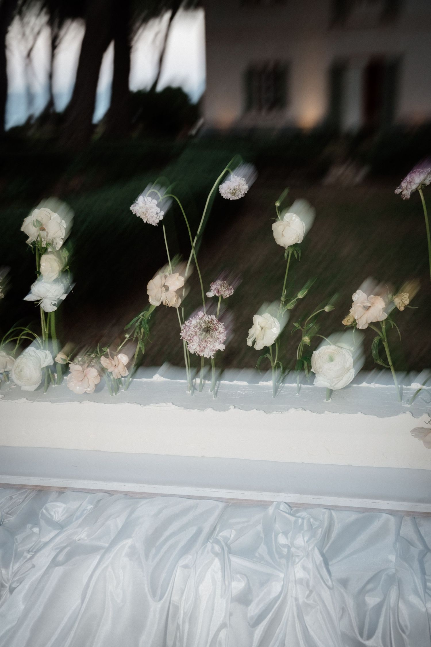 White flowers and alliums arranged along a draped table edge at an outdoor wedding reception.