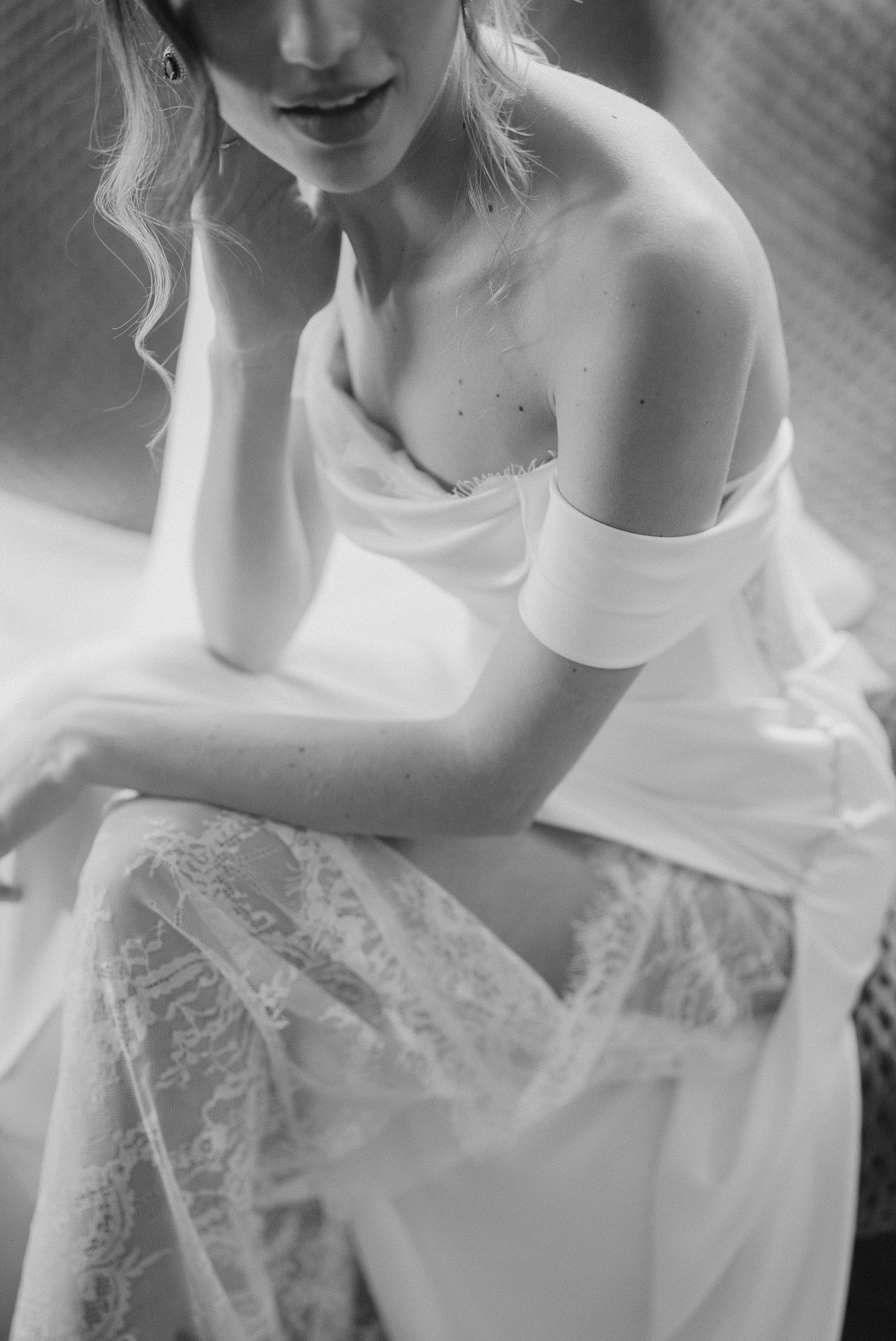 Bride in off-shoulder white gown with lace skirt sitting gracefully in black and white portrait.