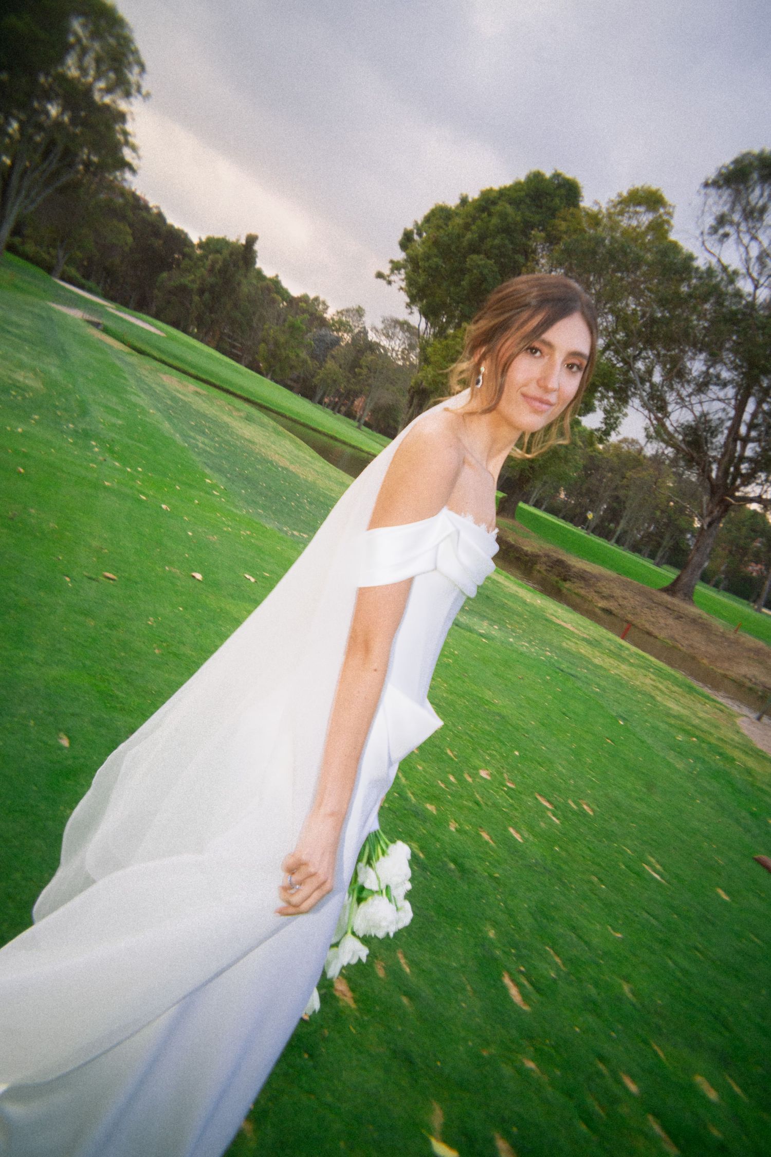 Bride in off-shoulder white gown holding roses on lush green lawn with trees in background.