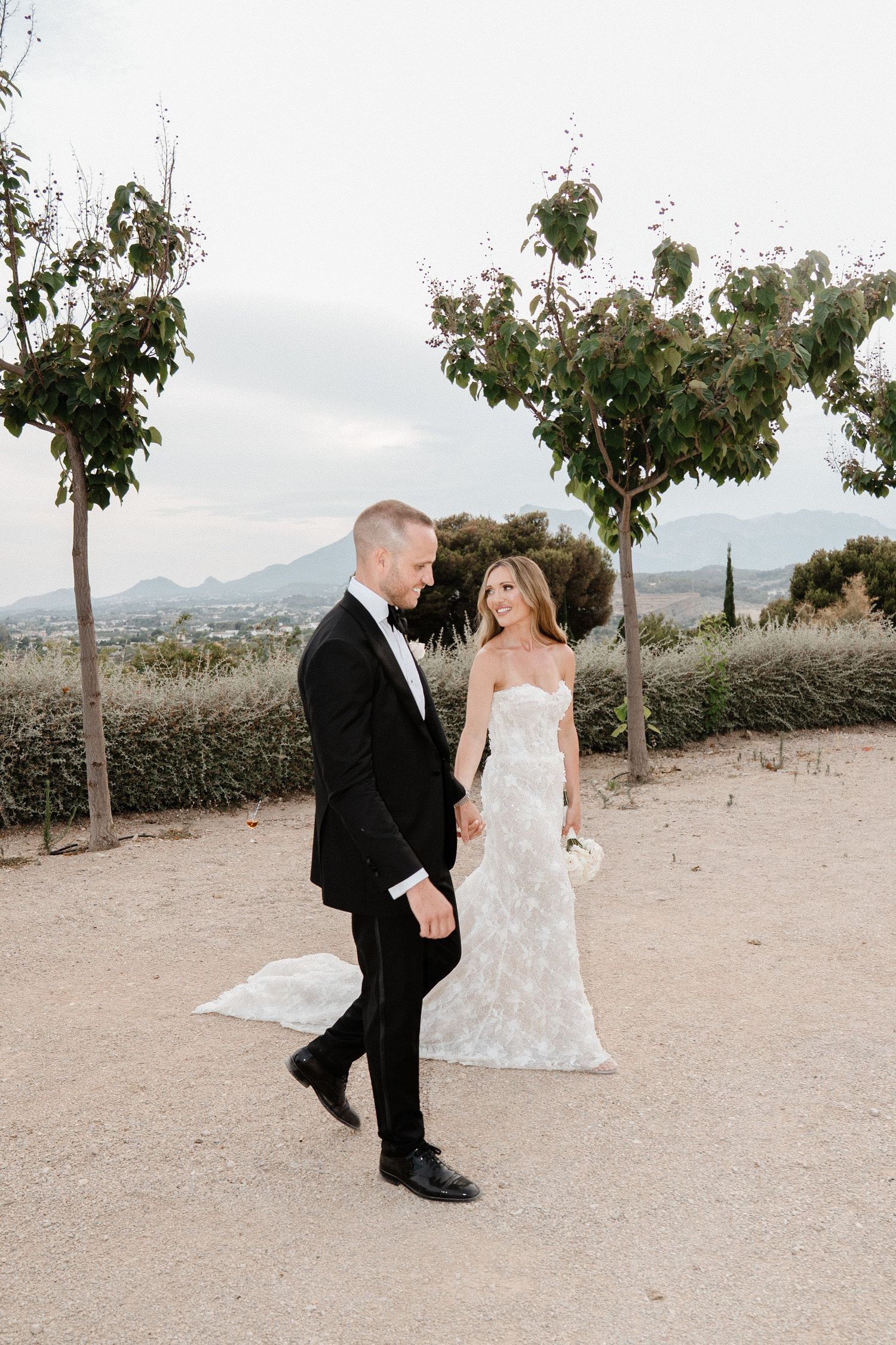 Bride and groom walking hand in hand through a scenic outdoor venue with mountains in the background.