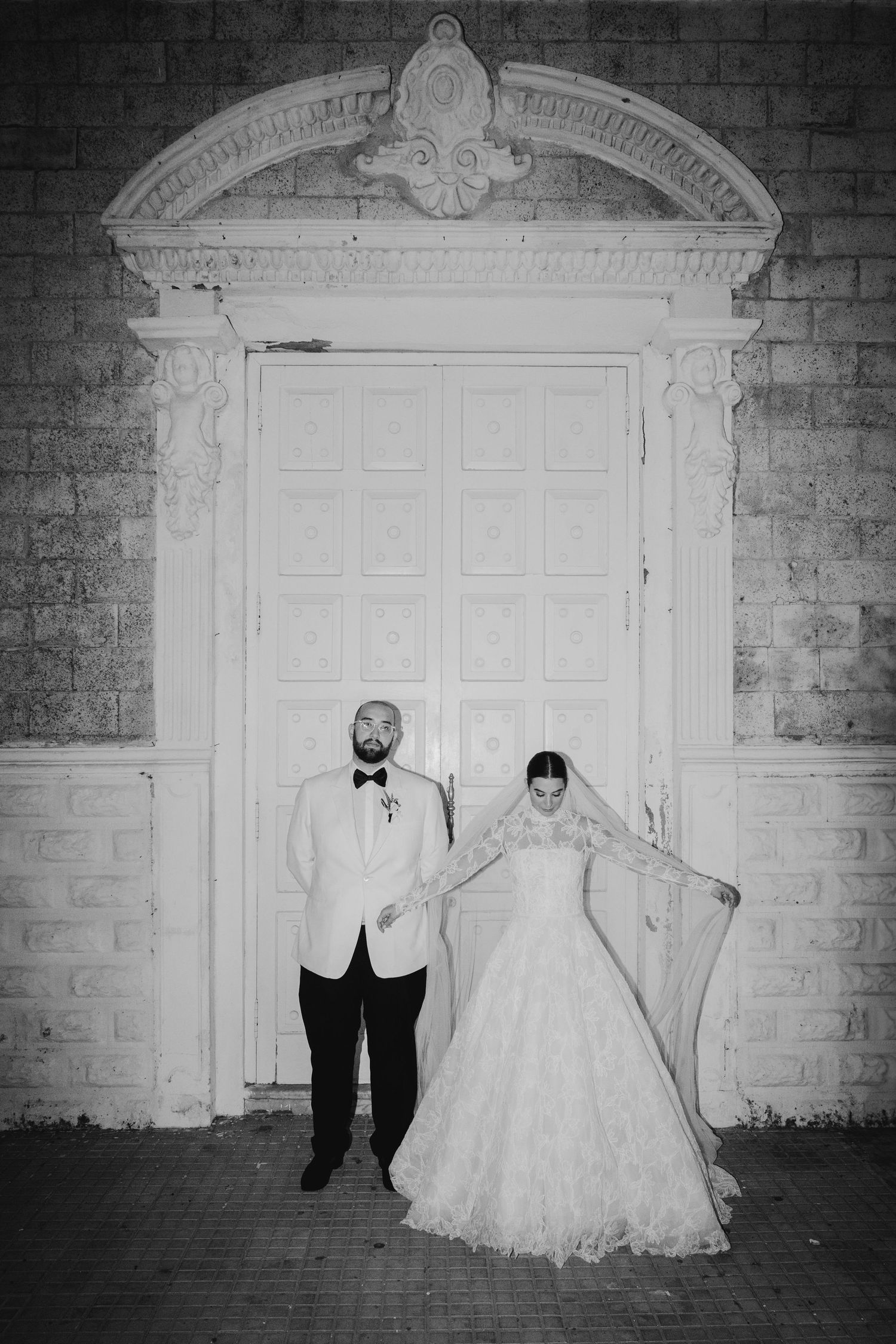 Bride in lace ballgown and bearded groom in white tuxedo pose before ornate white church doors.