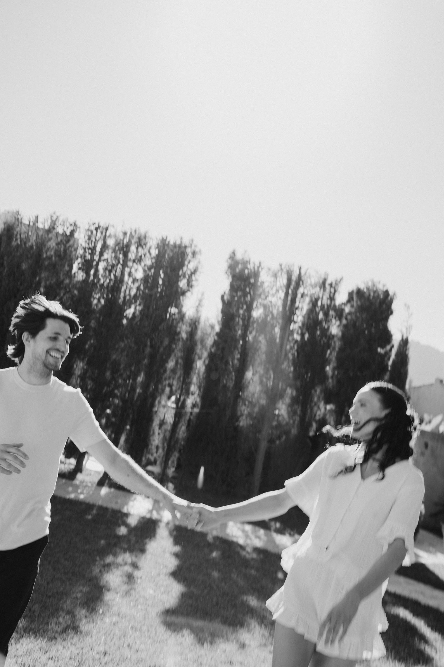 Black and white photo of a joyful couple holding hands and spinning outdoors with tall trees in the background.
