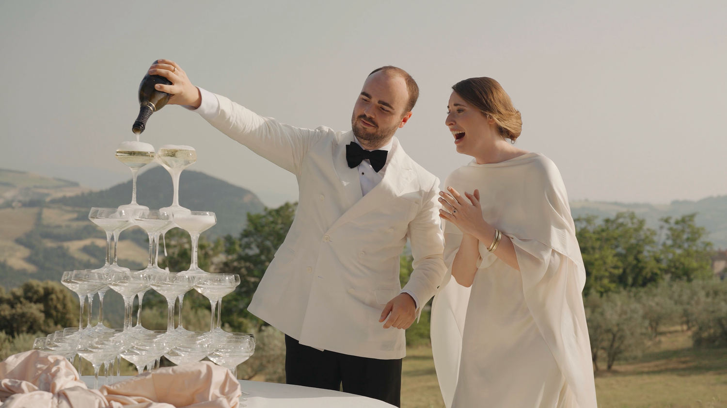 Bride and groom celebrating with a champagne tower pour against a scenic Tuscan countryside backdrop.