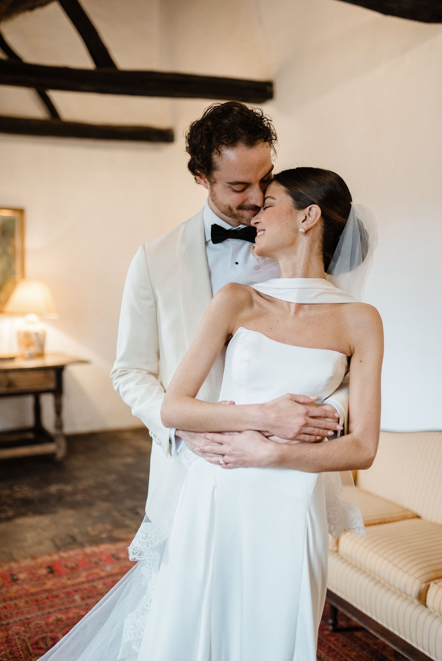 Groom in ivory tuxedo embraces bride in strapless gown in a warmly lit room.