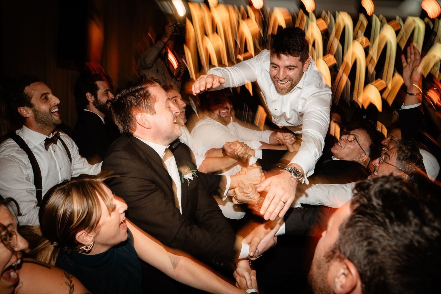 Groom lifted by cheering wedding guests on a lively dance floor during a festive wedding reception celebration.