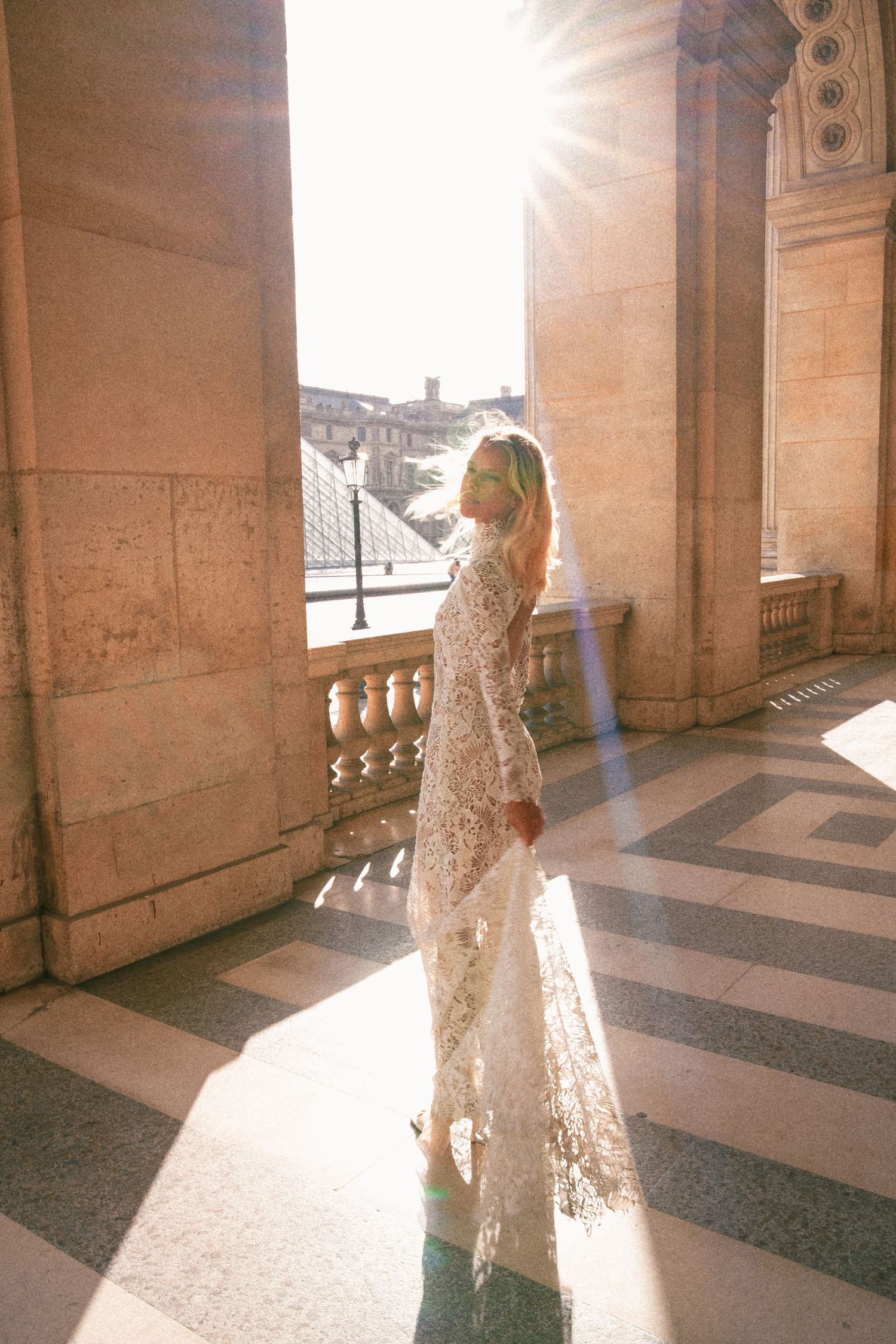 Woman in white lace gown standing in sunlit Louvre colonnade, Paris, with golden light streaming through stone columns.