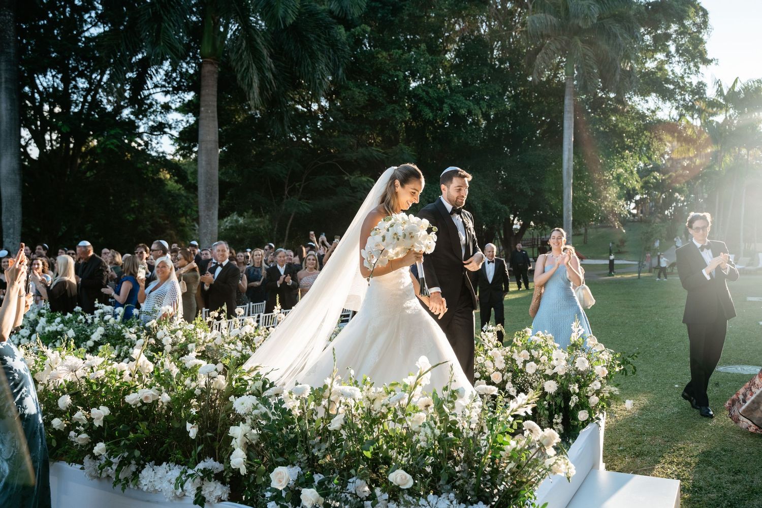 Bride and groom recessional through floral-lined aisle with veil flowing before cheering guests.